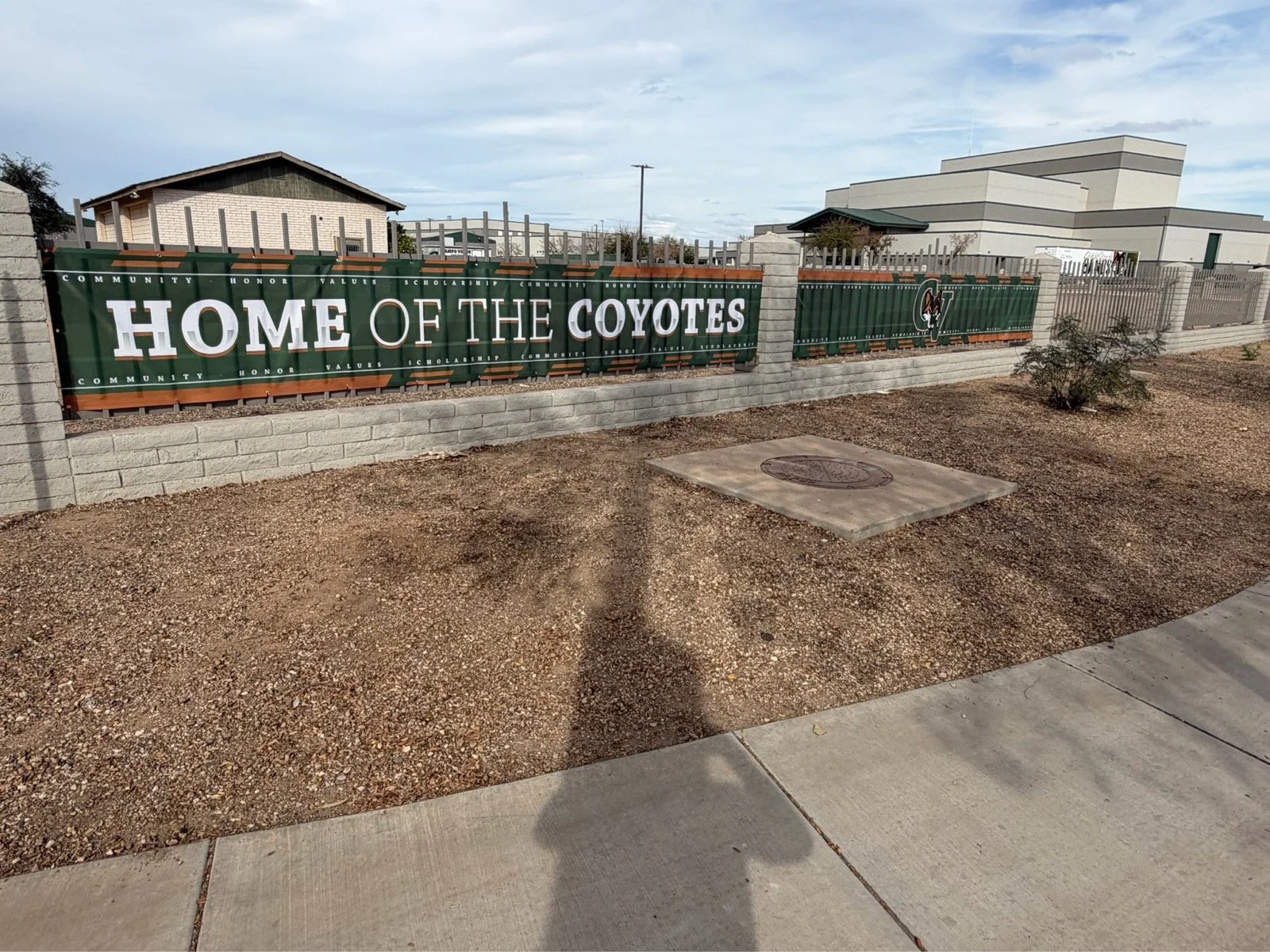 A green banner attached to a chain-link fence with a brick base, displaying the words 'HOME OF THE COYOTES' in large white letters, with smaller words 'COMMUNITY, HONOR, VALUES, SCHOLARSHIP' repeated along the top and bottom.