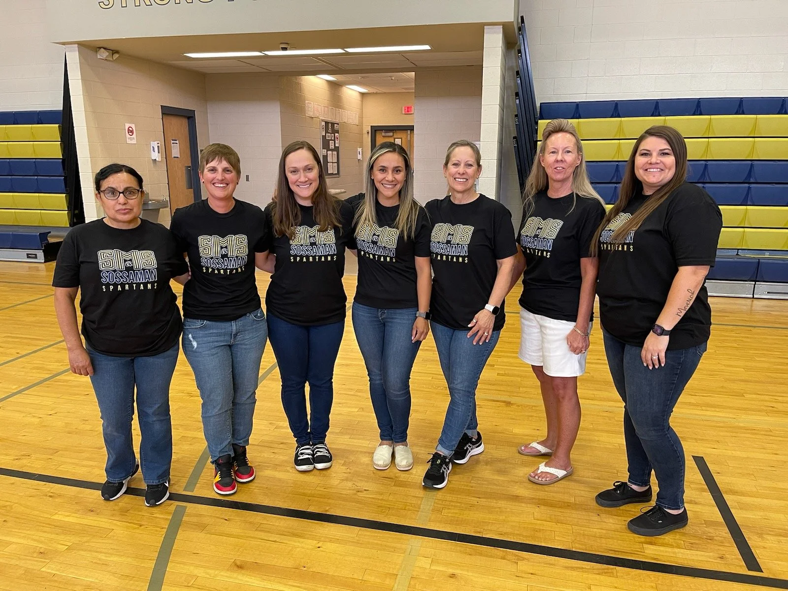 A group of seven women standing in a gymnasium, all wearing matching black t-shirts with white text that says 'SMS Sossaman Spartans,' posing for a photo.