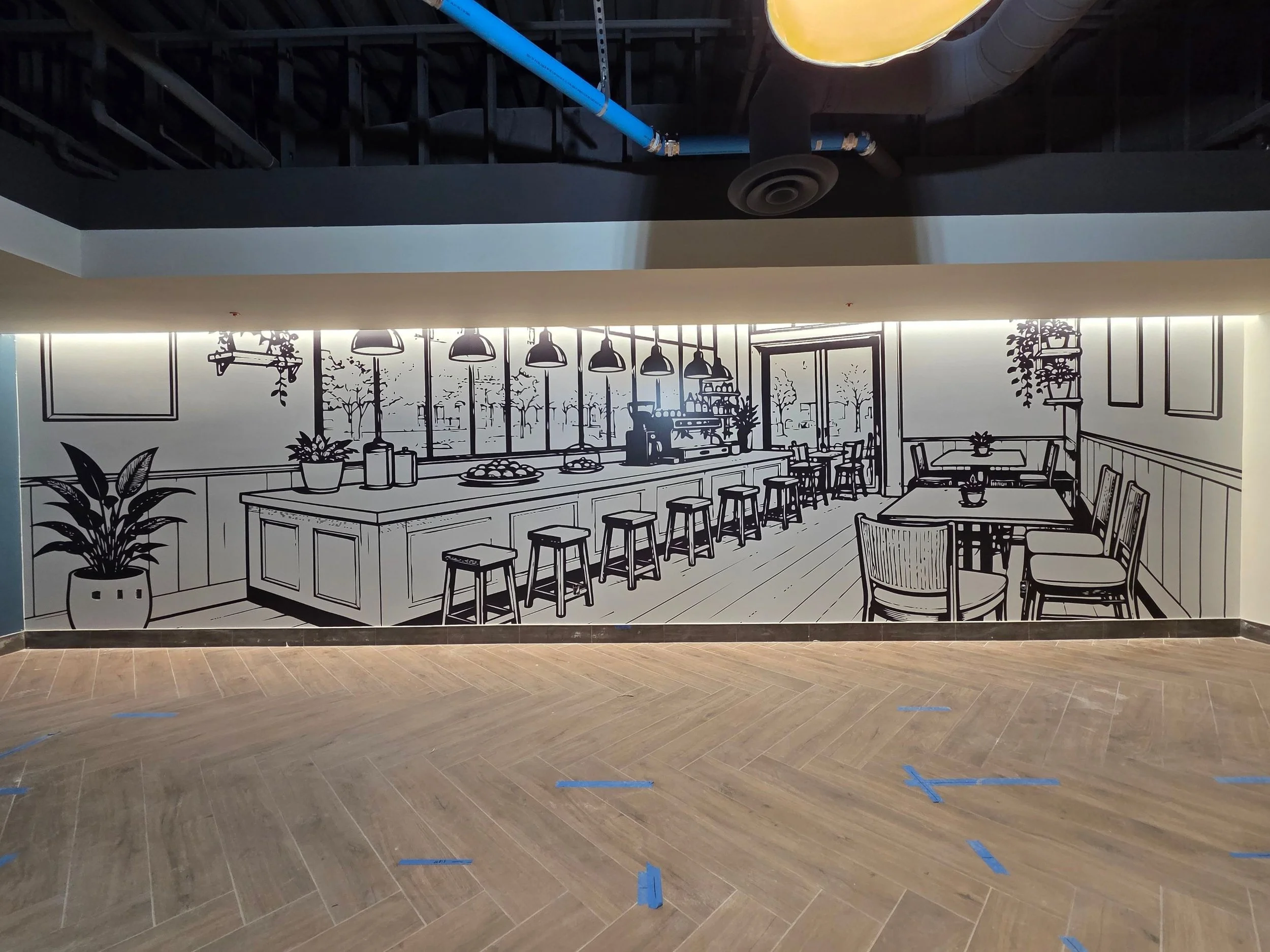 A wall mural of a black-and-white sketch of a cozy cafe interior with tables, chairs, hanging lights, plants, and a street view with trees outside. The mural is on a light-colored wall in a space with wood flooring.