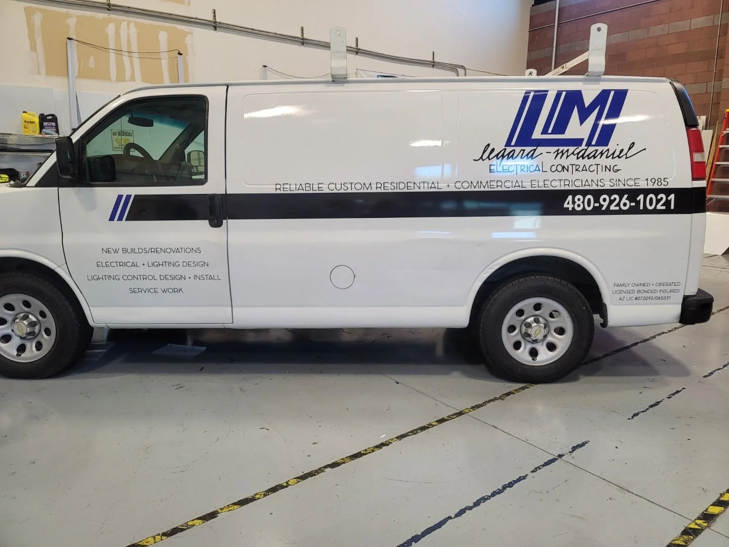 White company van with blue and black logo and text advertising electrical contracting services, parked inside a garage or warehouse with tools and supplies nearby.