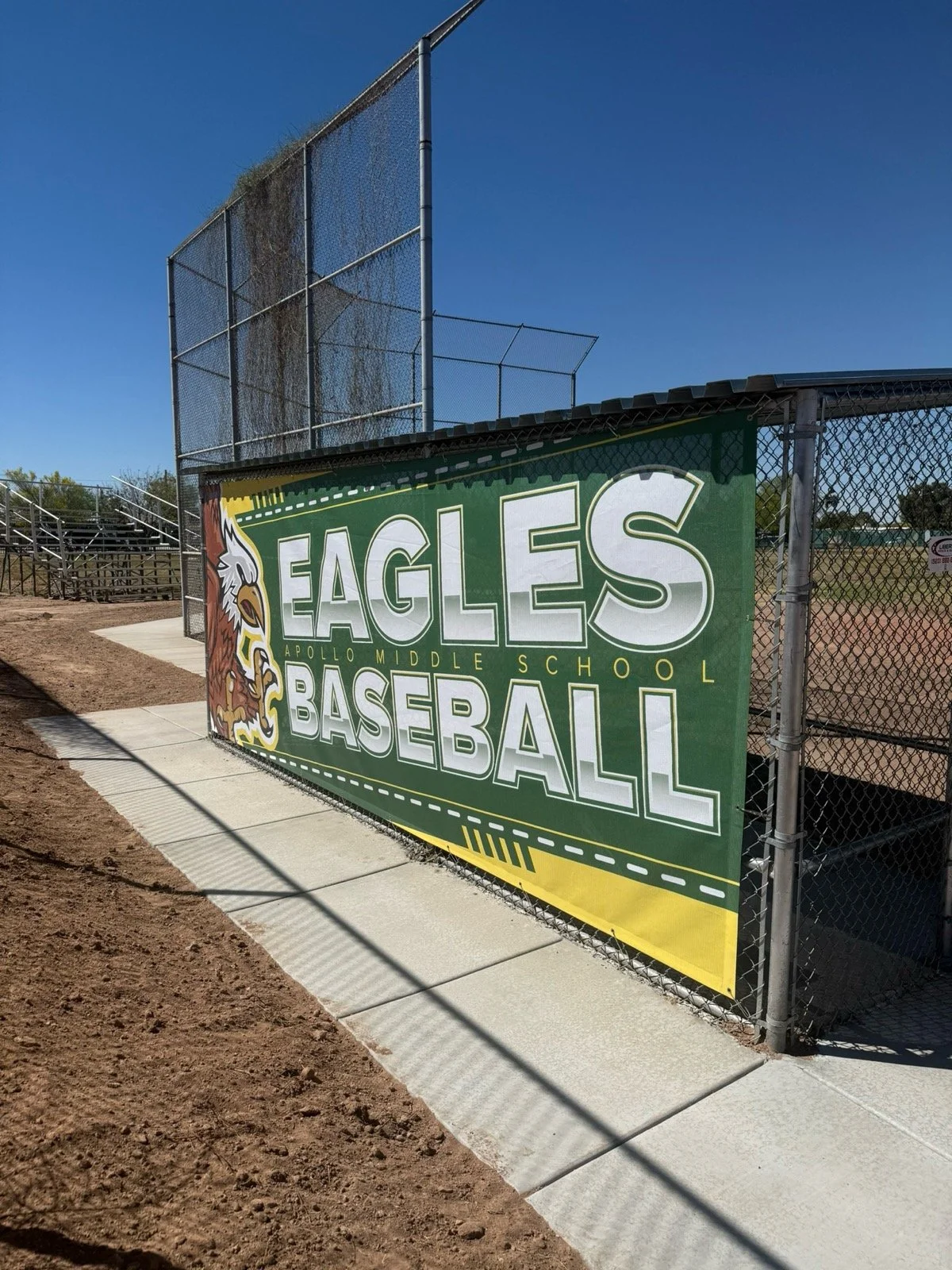 A green sign with the words "Eagles Baseball" and a logo of an eagle head, attached to a chain-link fence at a baseball field.