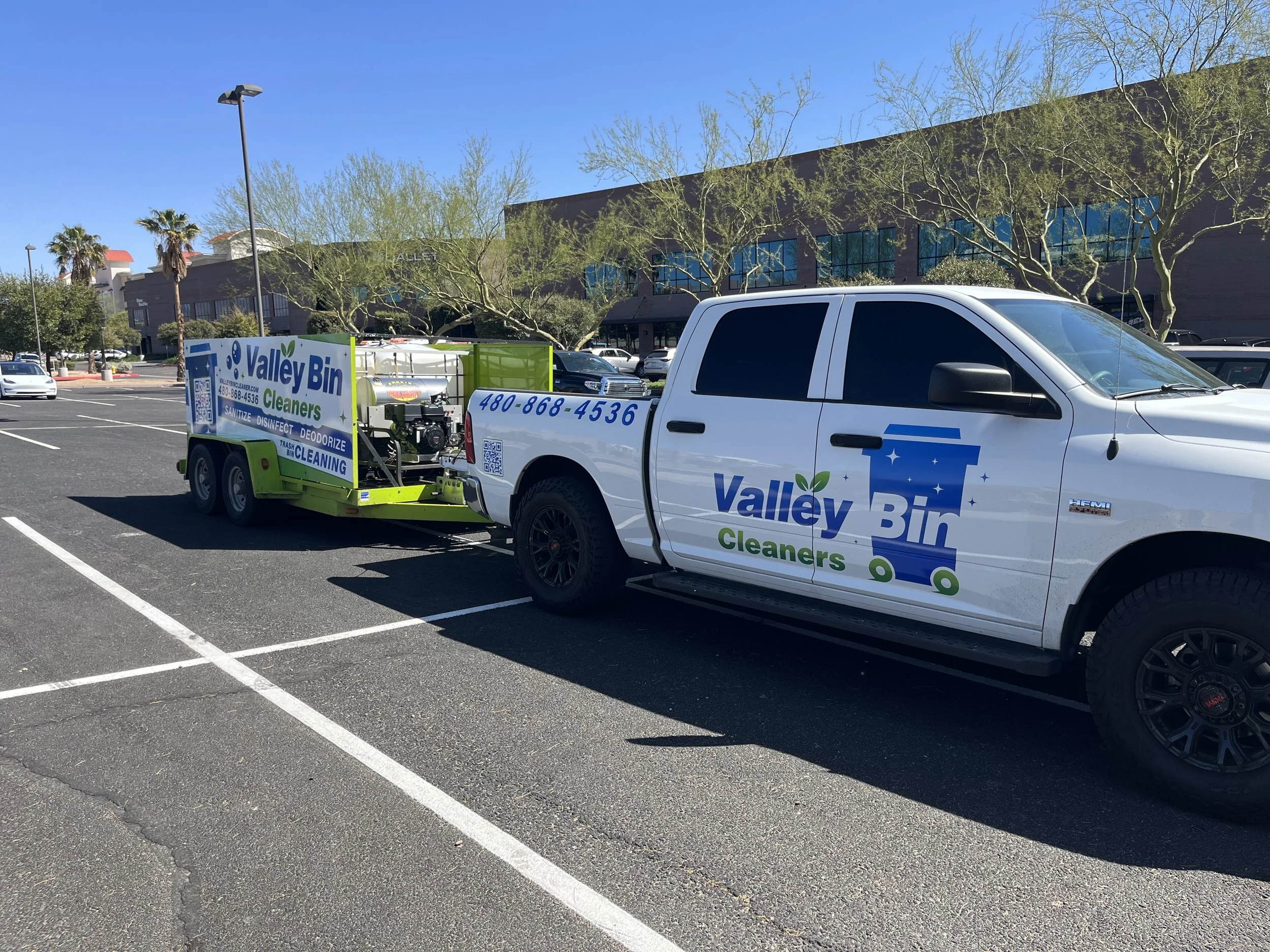 A pickup truck with Valley Bin Cleaners branding attached to a trailer, parked in a parking lot with several trees and a building in the background.