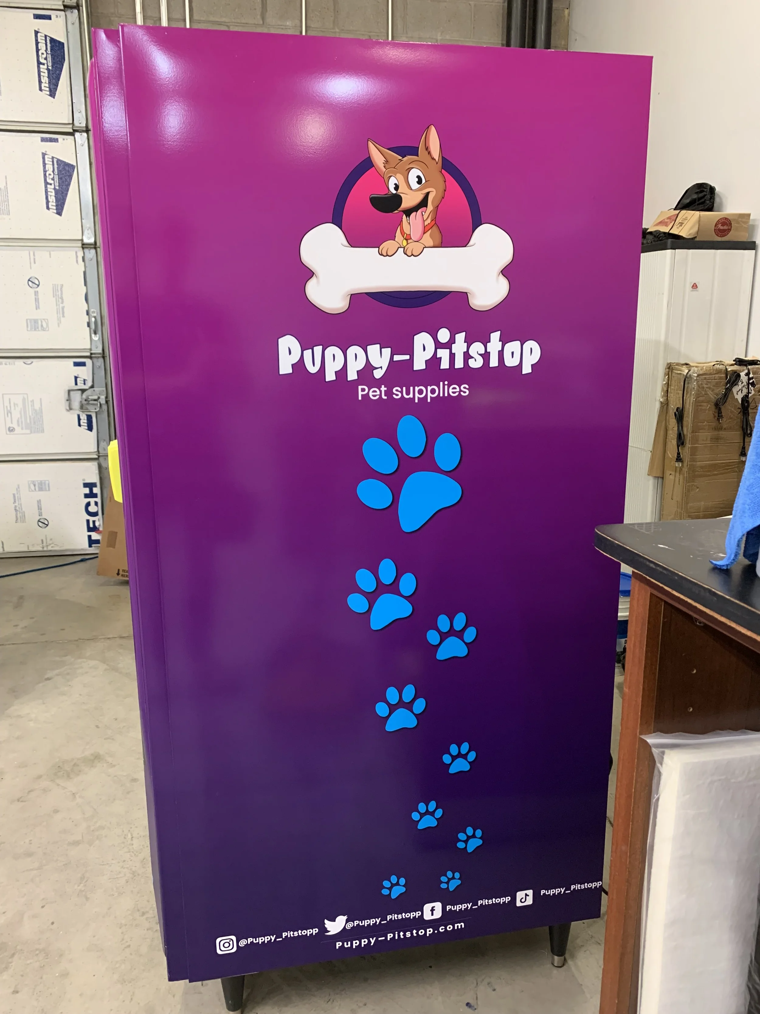 A purple sign with a cartoon dog smiling and sticking out its tongue, sitting behind a large bone. The sign displays the text 'Puppy-Pitstopp' and 'Pet supplies', along with blue paw prints. Social media icons and handles are at the bottom.