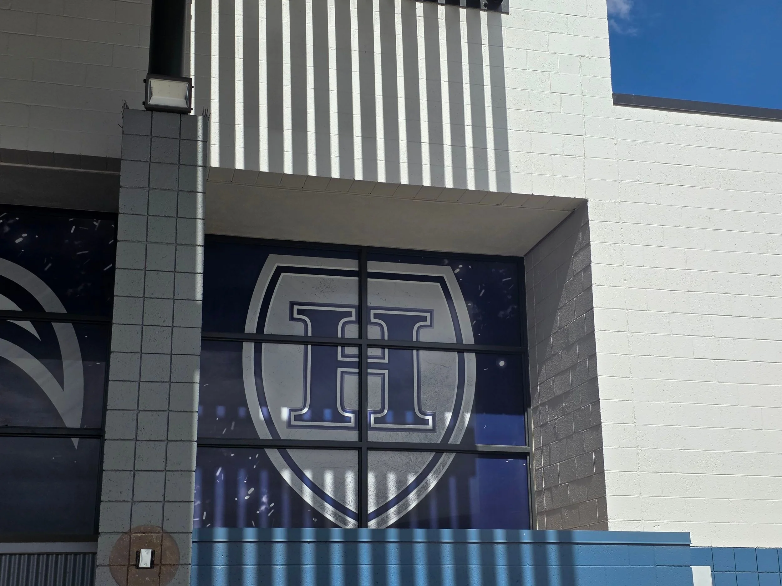 The image shows the exterior of a building with a white brick wall, a large sign with a shield logo and the number 25, and a black door. The sky is blue with some clouds, and there are grill openings on the wall and a light fixture on a post.
