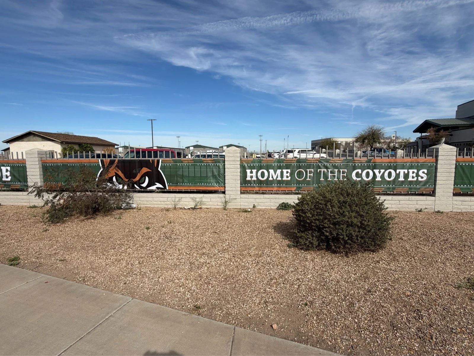 A fence with a green banner displaying a coyote mascot and the words "Home of the Coyotes" in front of a parking lot with cars and buildings, under a partly cloudy sky.