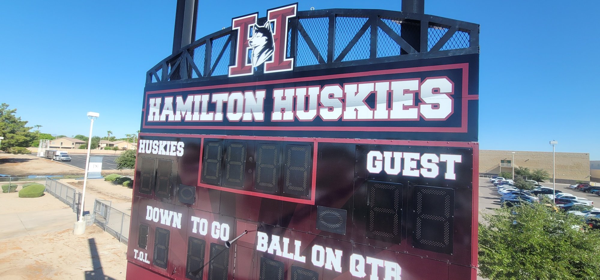 School football scoreboard displaying 'Hamilton Huskies' with a mascot of a husky dog, under a clear blue sky with a parking lot and school buildings in the background.