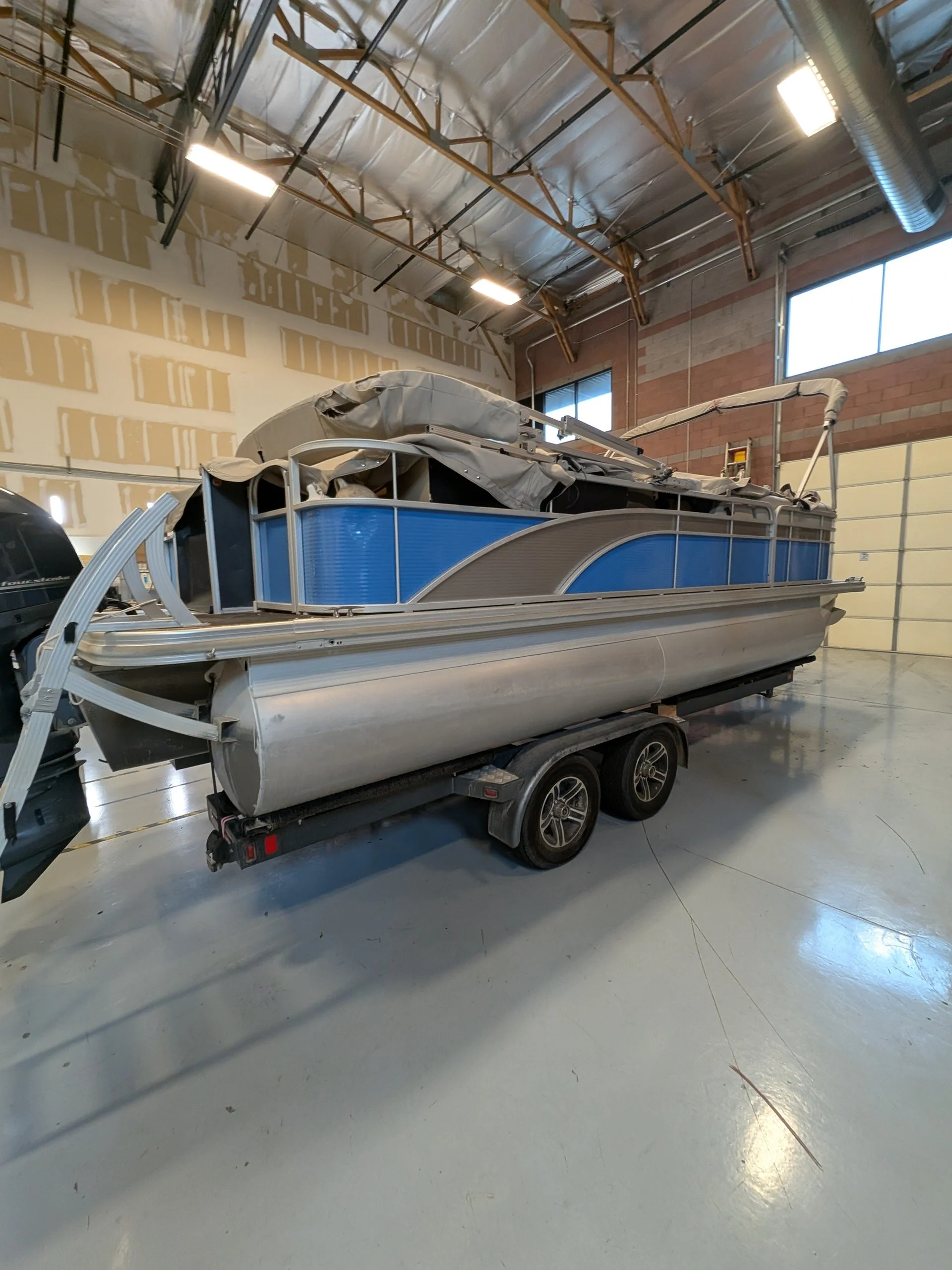 A pontoon boat with blue sides and a beige cover on the deck, parked inside a large unfinished garage with exposed insulation and walls, and a concrete floor.