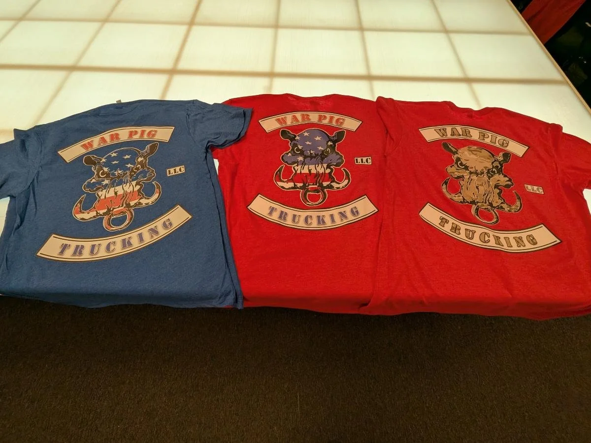 Three t-shirts with a graphic of a pig's head and the words "War Pig Trucking" on them, in blue, red, and tan colors.