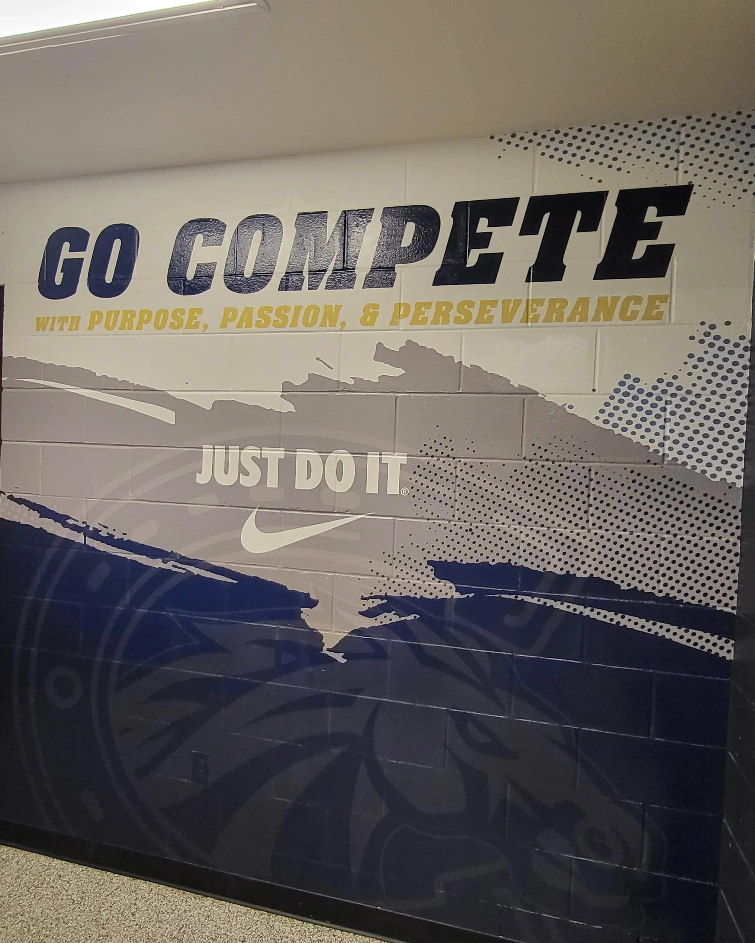 A wall with Nike branding, featuring the phrase 'GO COMPETE' in large bold letters, and the slogan 'WITH PURPOSE, PASSION, & PERSEVERANCE' underneath. The wall is decorated with stylized graphics of the United States map and Nike swoosh logos.