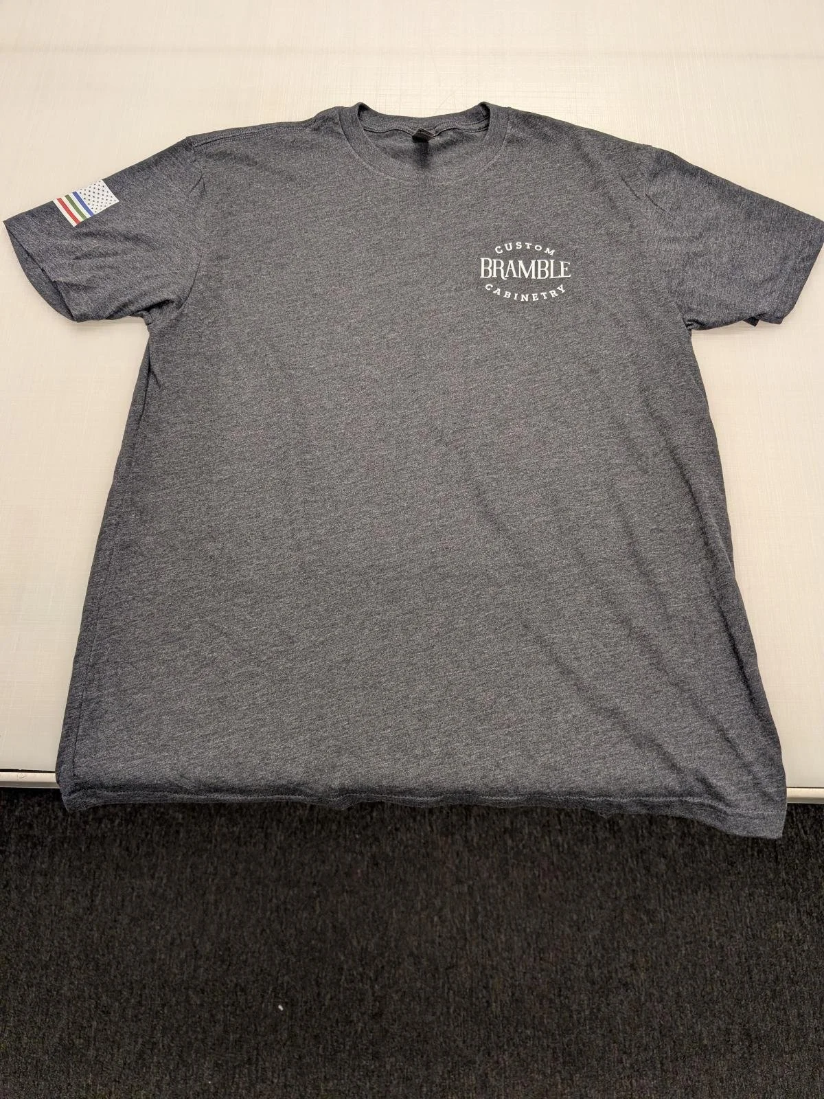 Gray T-shirt with a small logo on the chest reading "Custom Bramble Cabinetry" and a small, colorful striped patch on the left sleeve.