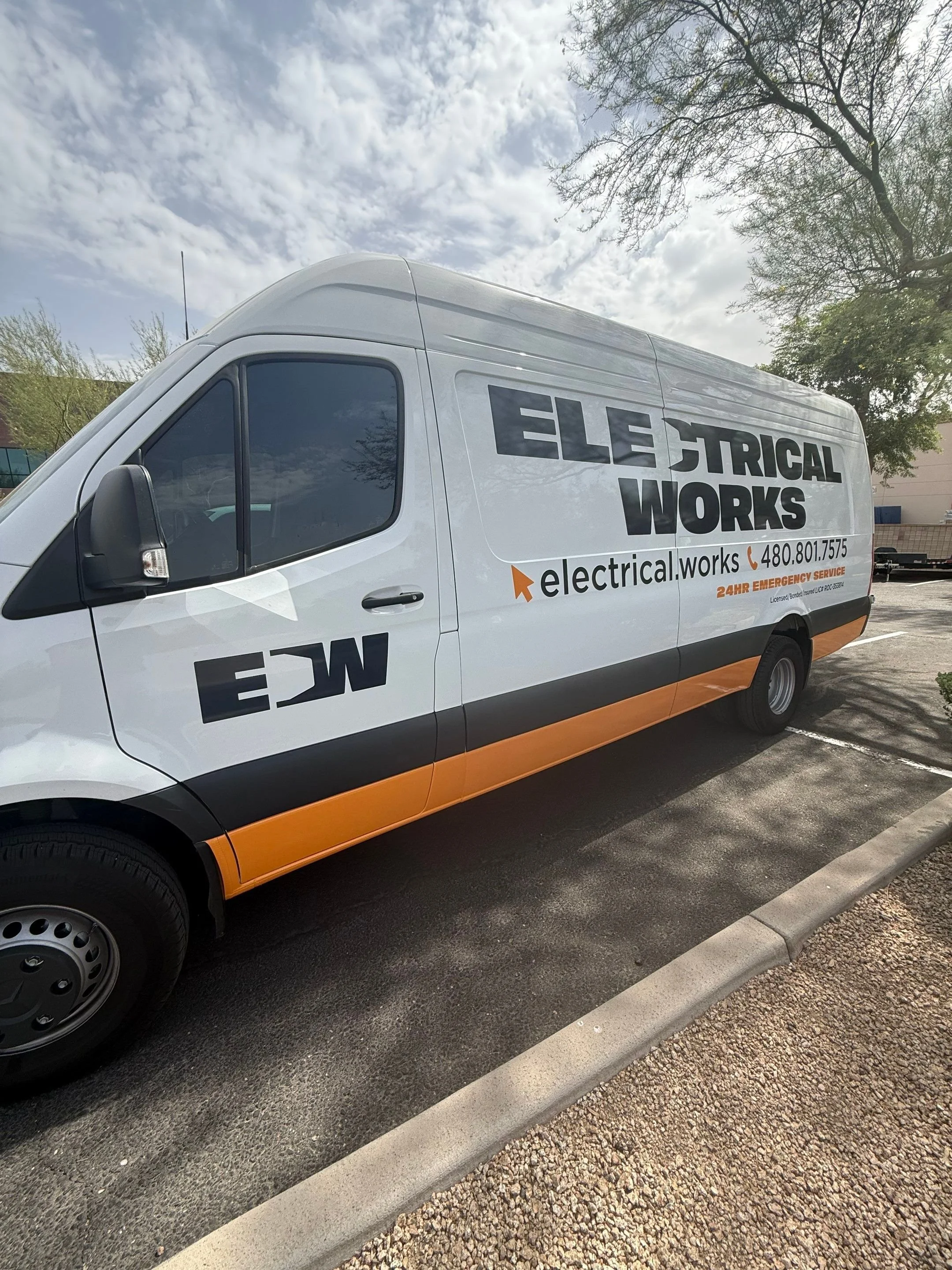 A white service van for electrical work is parked in a parking lot under partly cloudy sky, with trees and buildings in the background.
