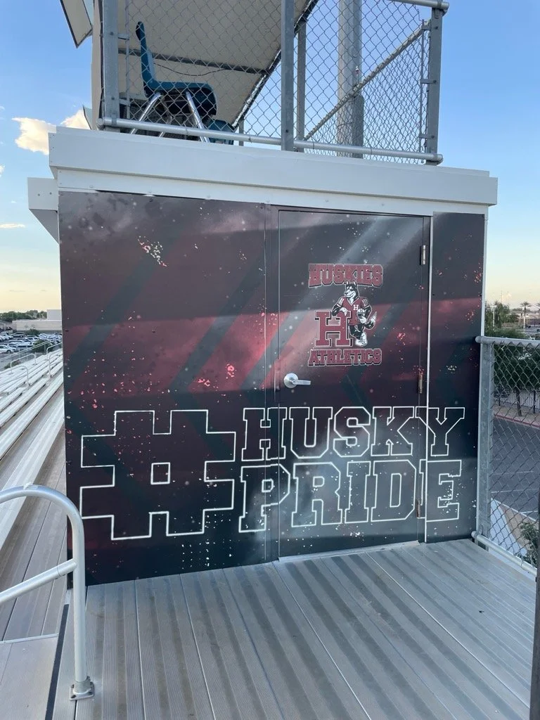 Decorative wall with the words 'Husky Pride' and a husky mascot logo for Husky Athletics, located on a sports stadium balcony or press box with fencing and a view of parking lot and sky.