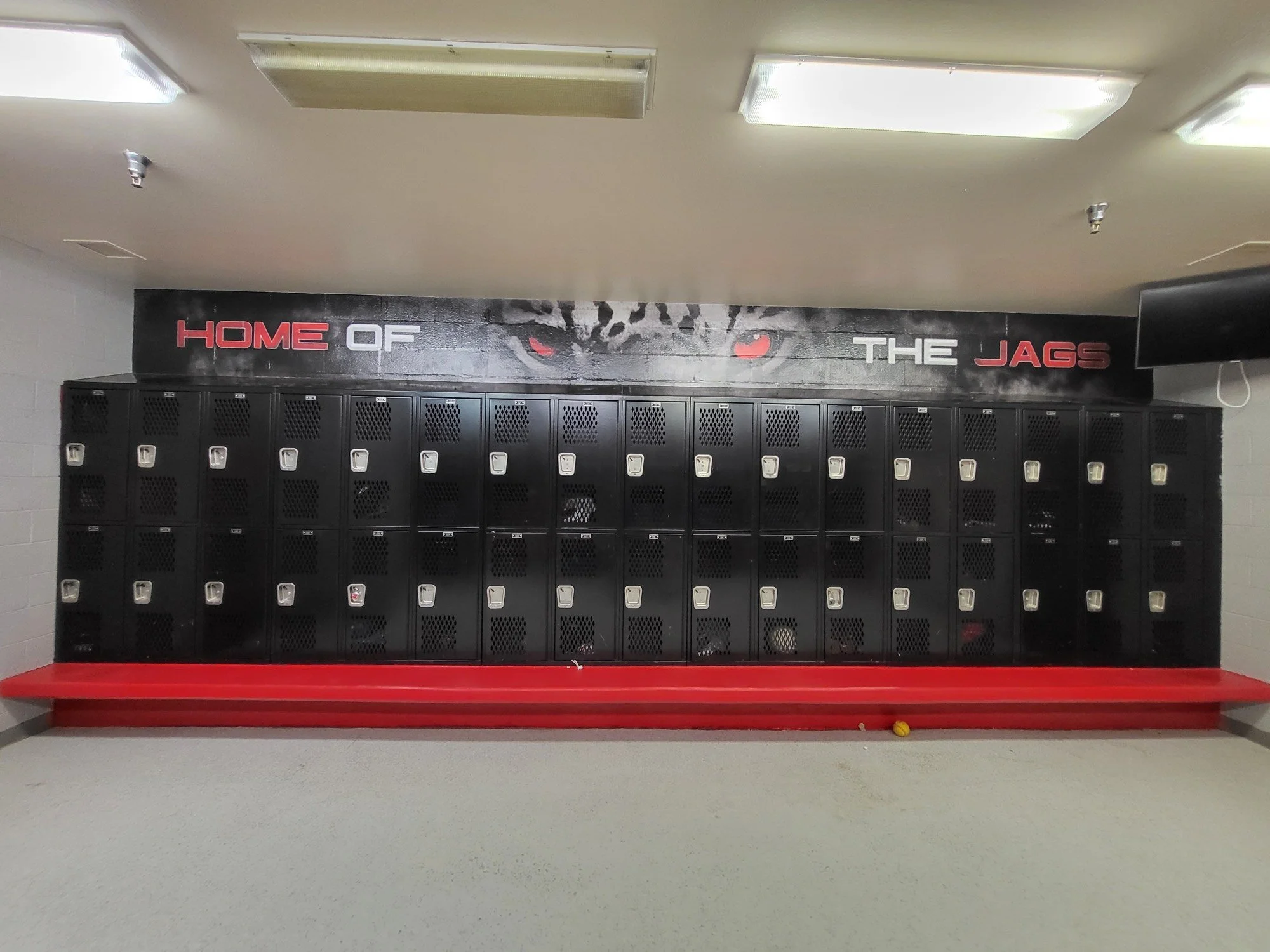 School locker room with black lockers, a red bench, a yellow tennis ball on the floor, and a wall decal reading 'Home of the Jags' with a jaguar's eyes.