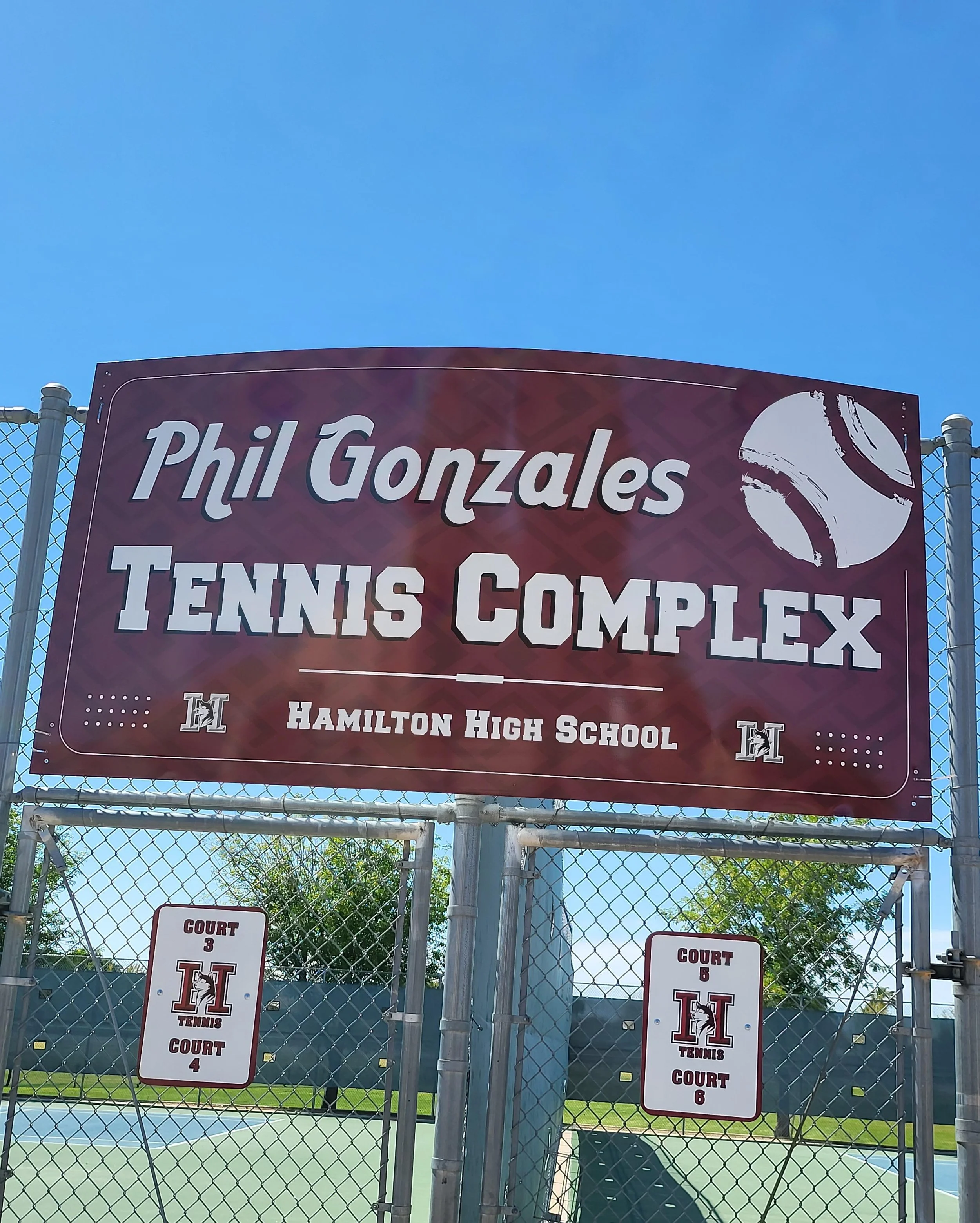Sign on a chain-link fence for the Hamilton High School tennis courts, with court numbers 3, 4, 6, and 8 visible underneath a large sign with the school name and a tennis ball logo. Blue sky in background.