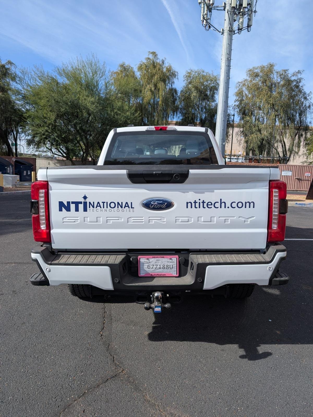 The rear of a white Ford Super Duty pickup truck parked in a parking lot, with decals for NTI National Technologies, ntech.com, and a pink AutoNation license plate.