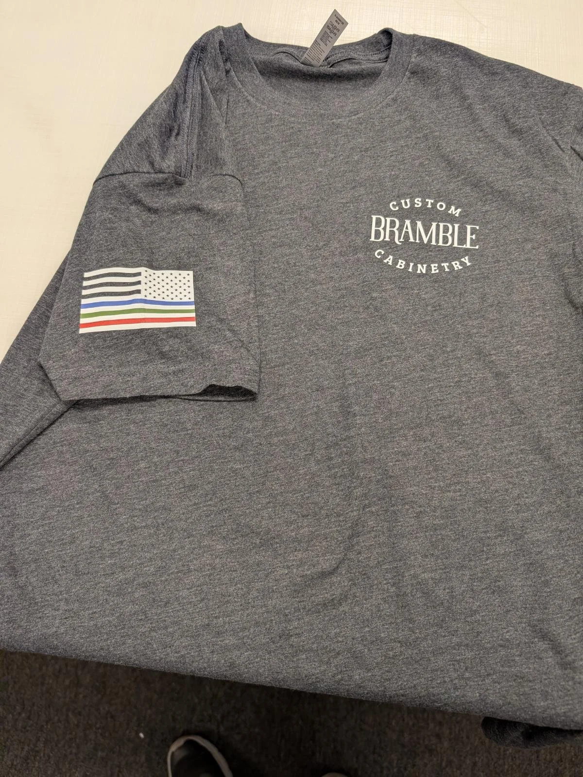 A gray T-shirt with a printed American flag with a thin blue line and a red stripe on the sleeve, and the words "Custom Bramble Cabinetry" printed on the front.