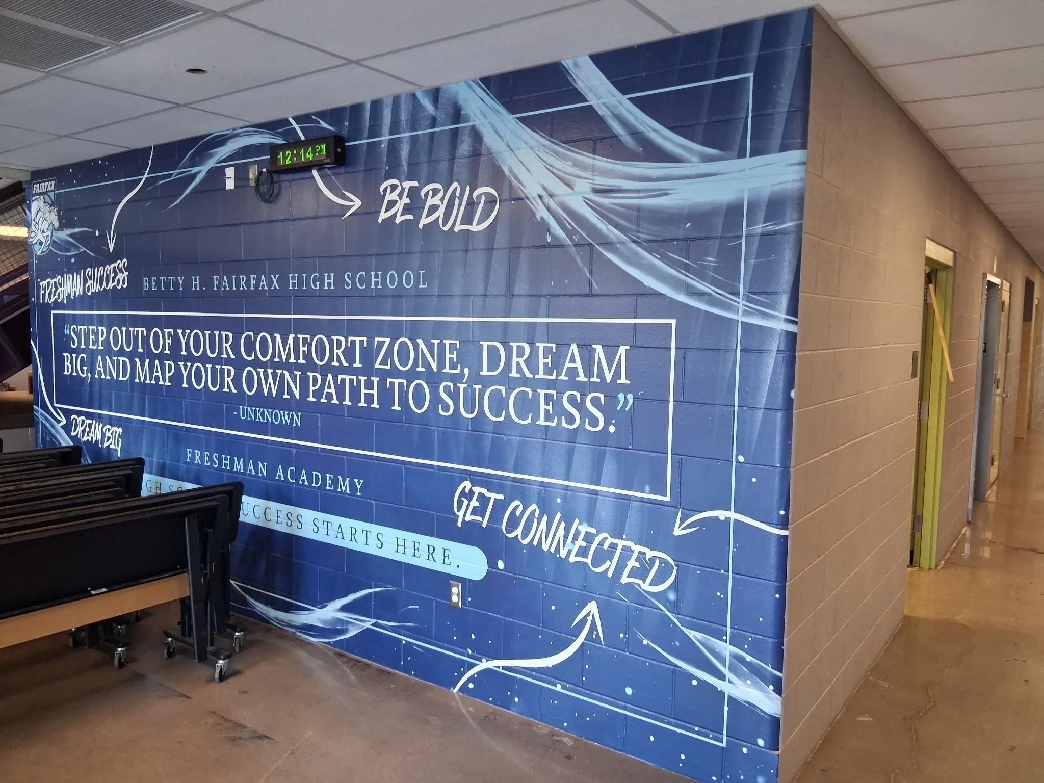 Blue school banner with inspirational quote, 'Step out of your comfort zone, dream big, and map your own path to success,' displayed inside a school corridor. The banner includes the school's name, Betty H. Fairfax High School, and has a digital cloc