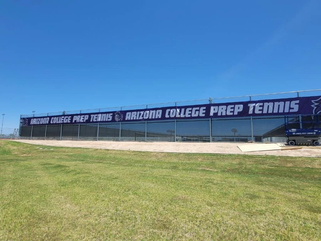 A large banner on a fence reads 'Arizona College Prep Tennis' with a purple background and white text, located at a tennis facility with a grassy field in the foreground and a clear blue sky above.