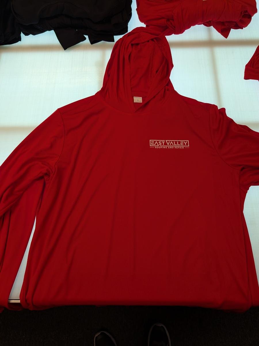 Red hoodie with a logo that reads 'East Valley Roofing and Repair' lying flat on a surface, with other pieces of clothing partially visible in the background.