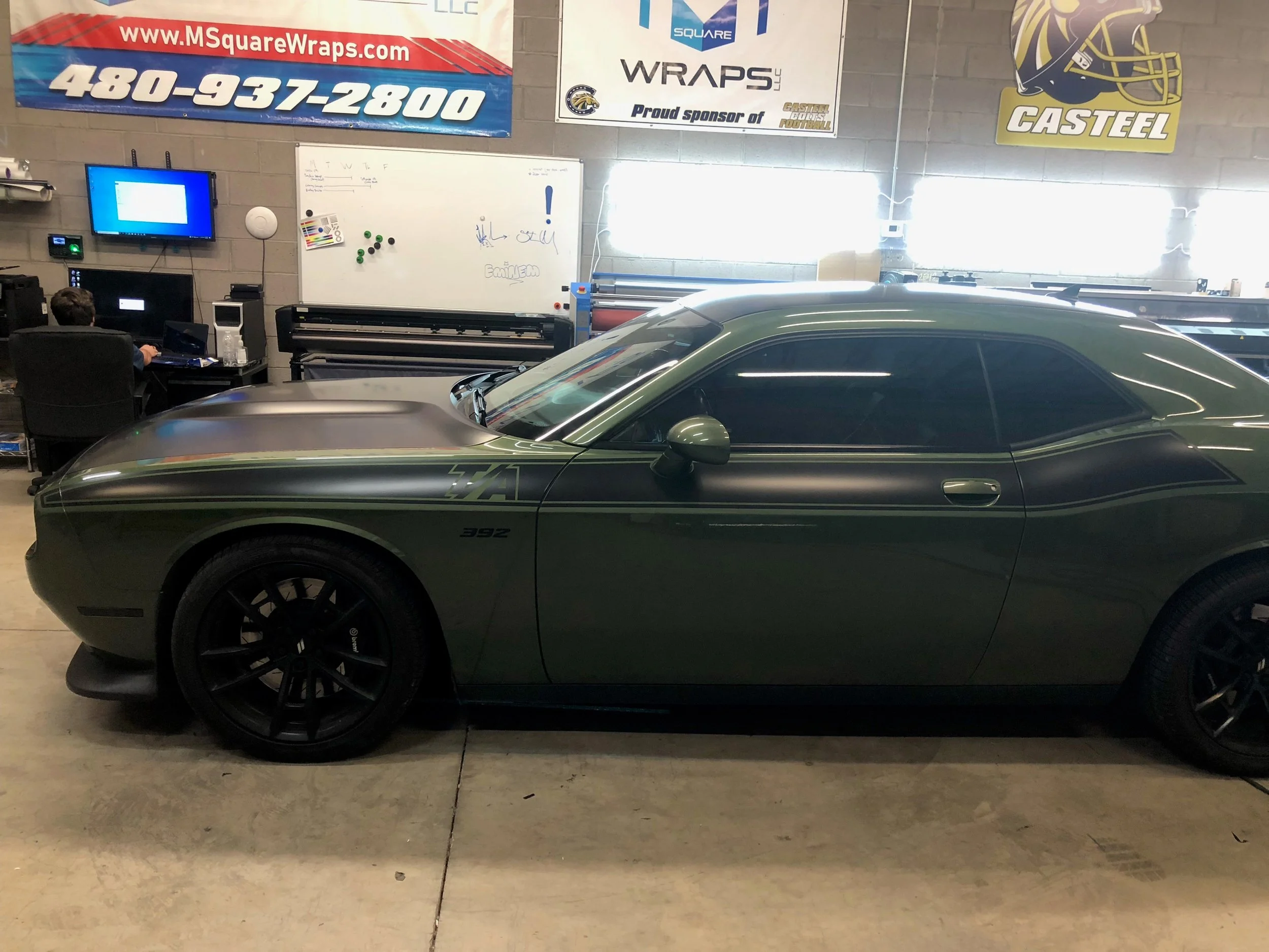 A gray sports car parked inside a workshop with banners on the wall, including a website, phone number, and logos for Wraps LLC and Castiel.