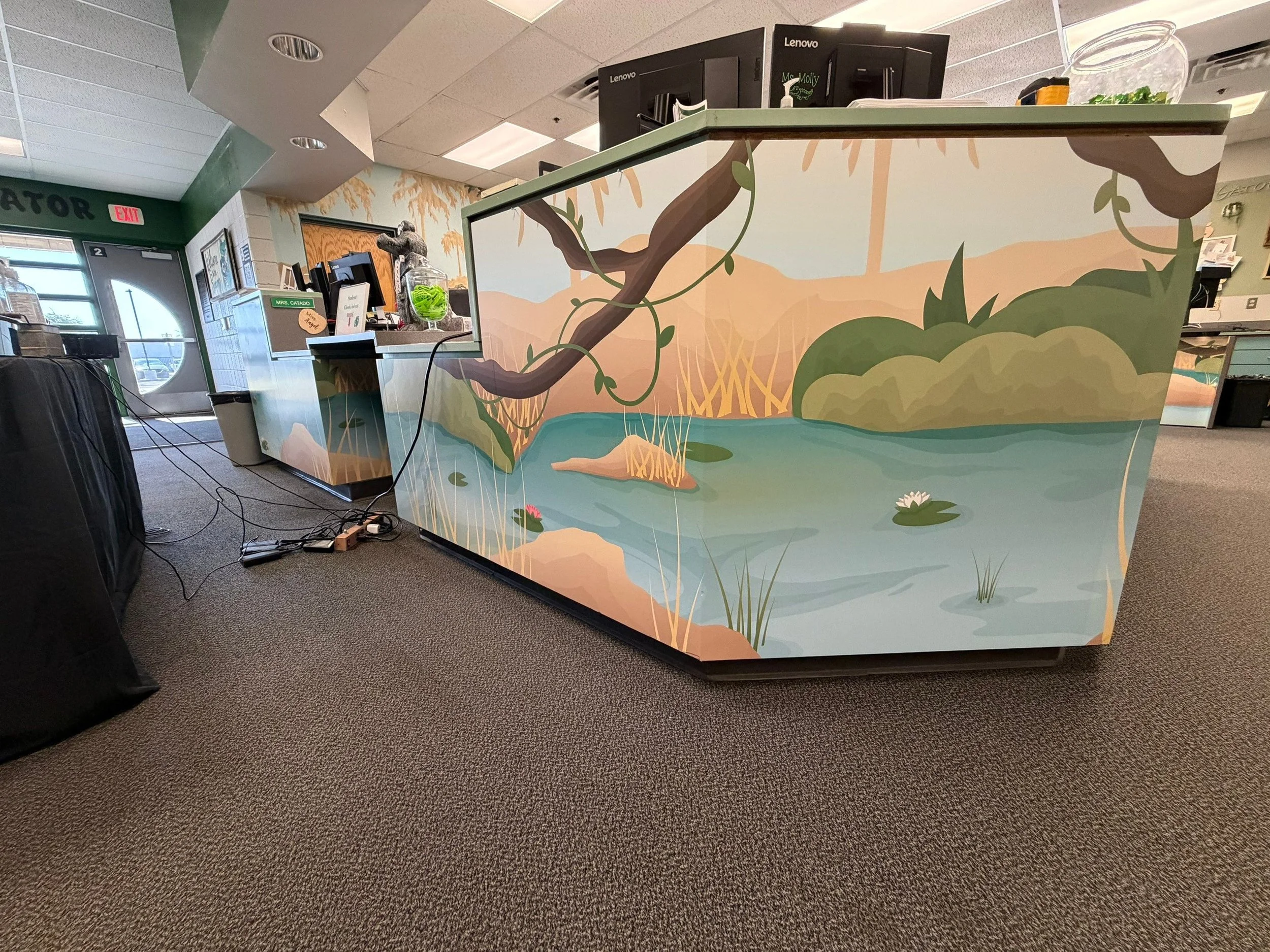 Interior of a cafe or restaurant featuring a colorful mural of a nature scene with water, rocks, and plants on the front desk, with surveillance monitors, computer equipment, and miscellaneous items on top.