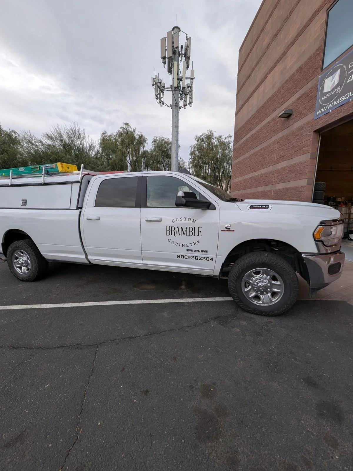 White work pickup truck with logo and text on the side, parked in front of a brick building and a cell tower under a cloudy sky.