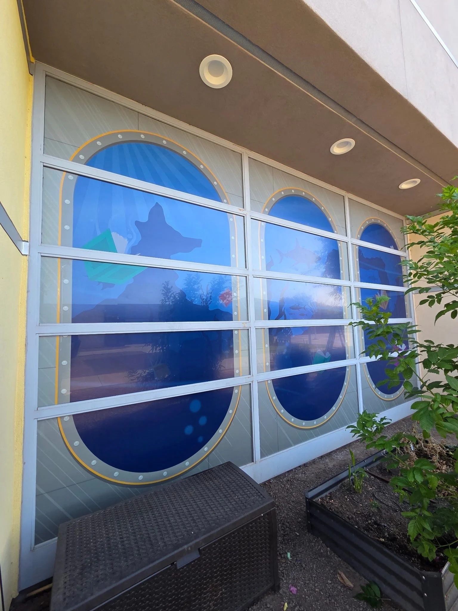 Exterior of a building with large window decals of fish and aquatic scenery, a small outdoor seating with black wicker tables and benches, and some potted plants.