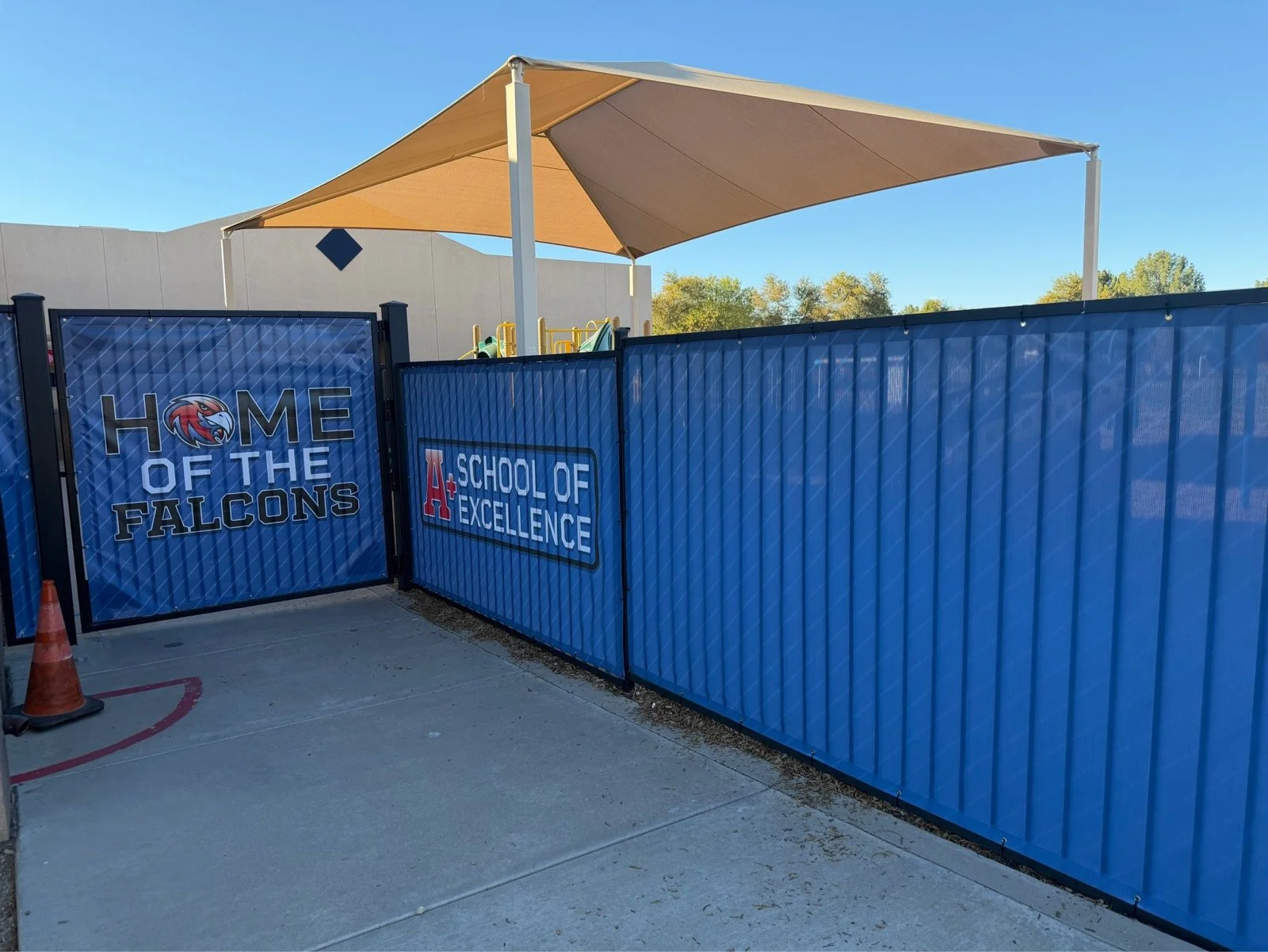 School playground entrance with signs that read "Home of the Falcons" and "A+ School of Excellence," blue metal fence and a shaded area with a beige canopy.