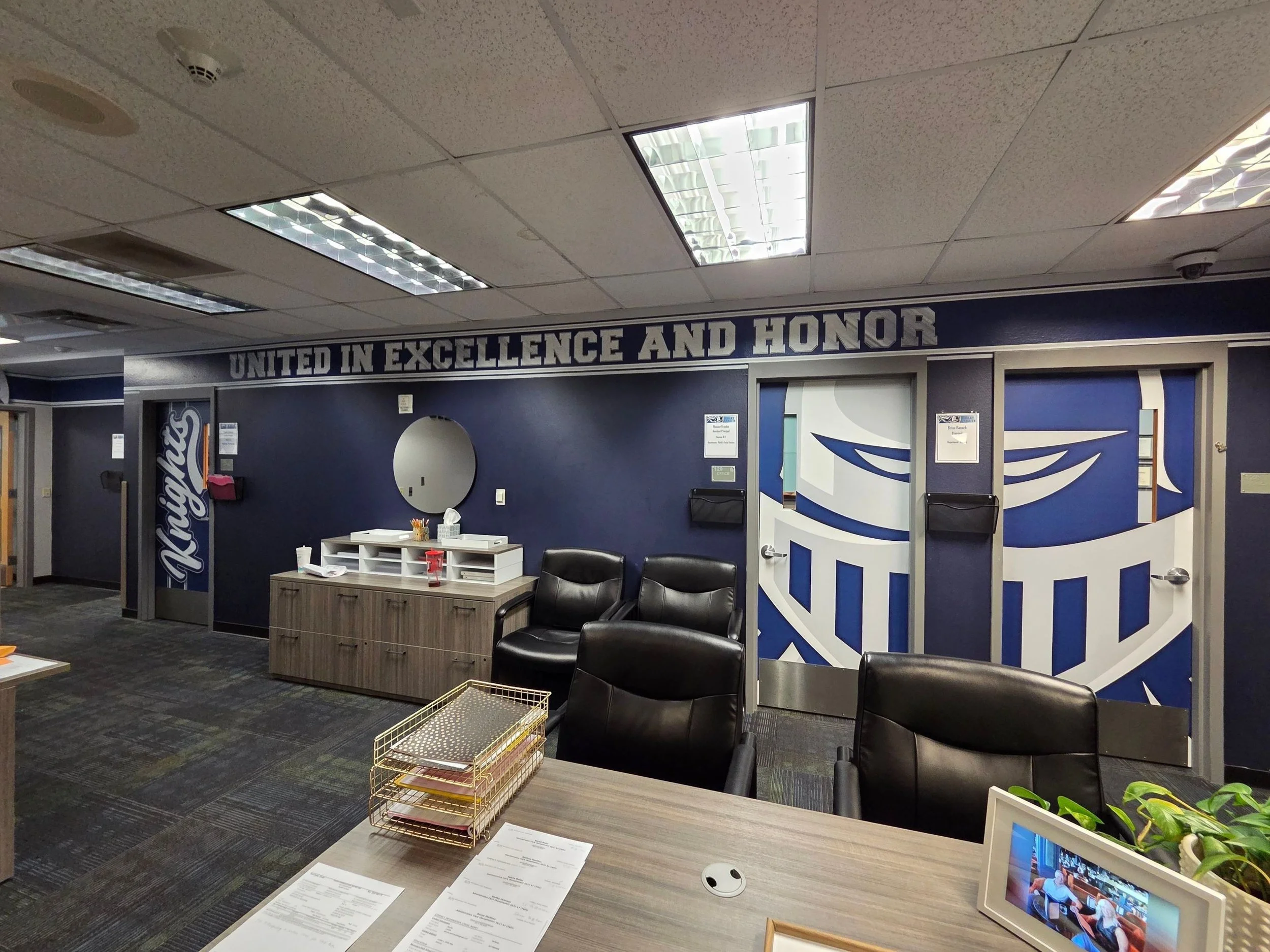 Office waiting area with chairs, a table with papers, and a wall with the phrase 'United in Excellence and Honor' in large white letters. There is a small monitor displaying a meeting or presentation, and some decorative plants.