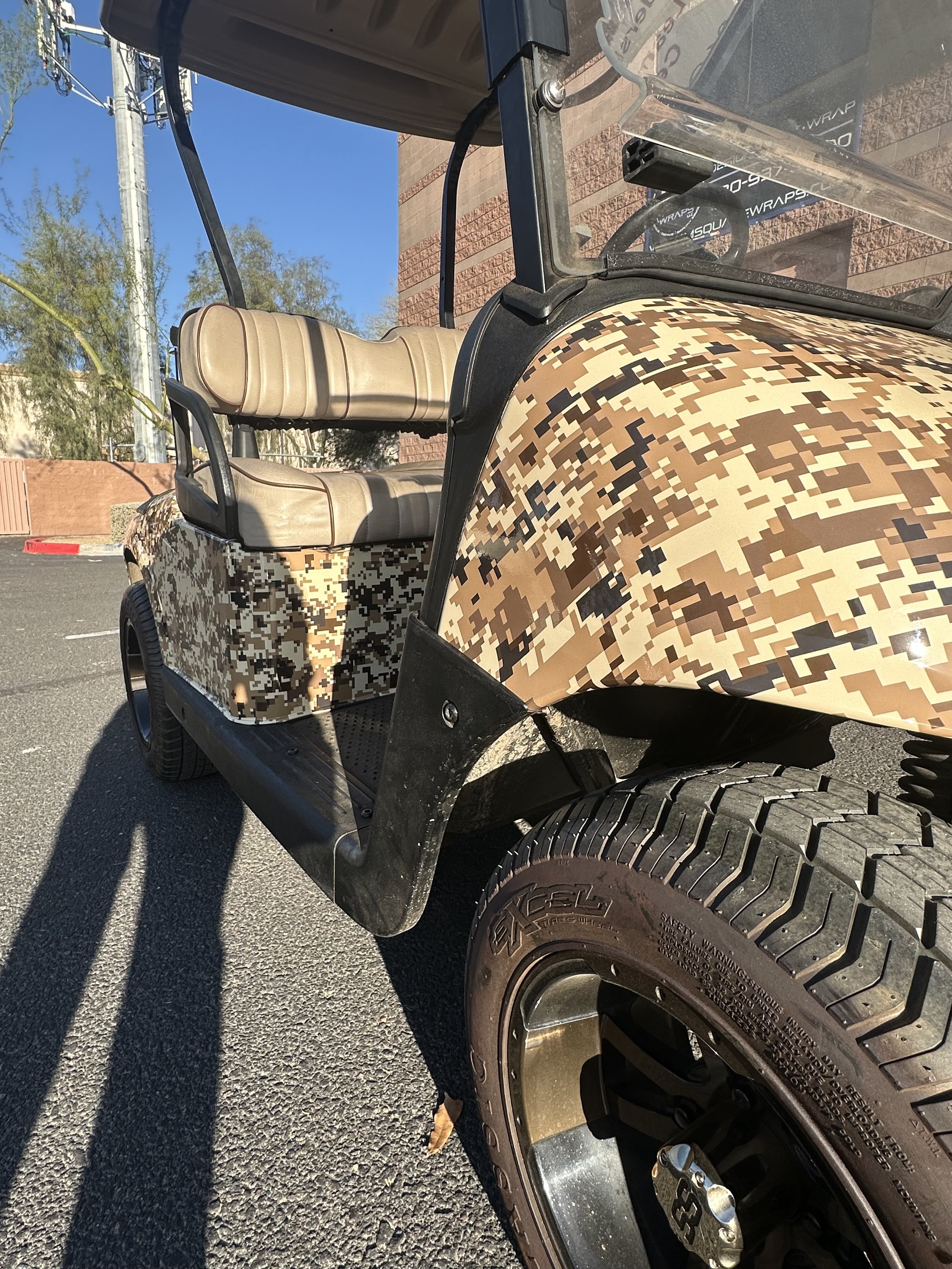 A golf cart with digital camouflage pattern on the body, beige seats, and large off-road tires parked on a paved parking lot next to a brick wall.