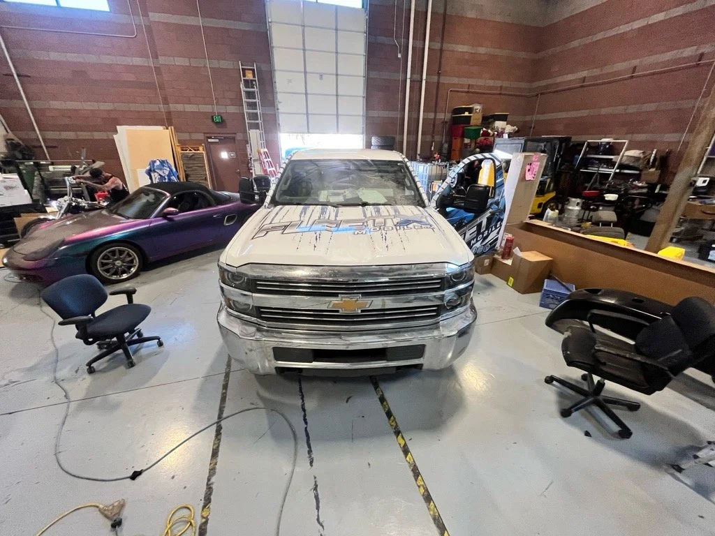 Indoor warehouse with a white Chevrolet truck in the center, a purple sports car to the left, and a partially visible black and yellow vehicle to the right, surrounded by chairs, boxes, shelves, and storage items.