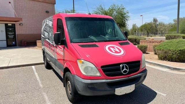 Red Mercedes-Benz van with N logo parked in a parking lot near a building and greenery.