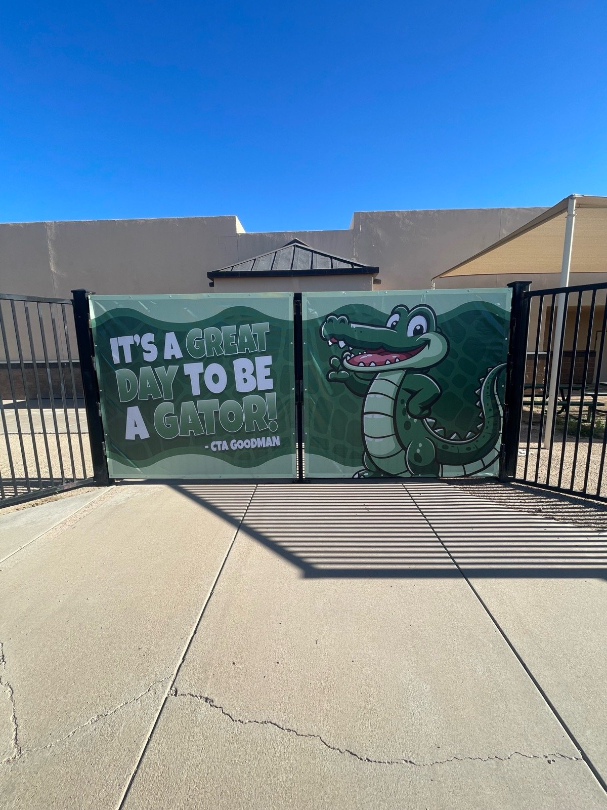 A gate with a cartoon crocodile and a sign that says "It's a great day to be a gator!" under a clear blue sky.
