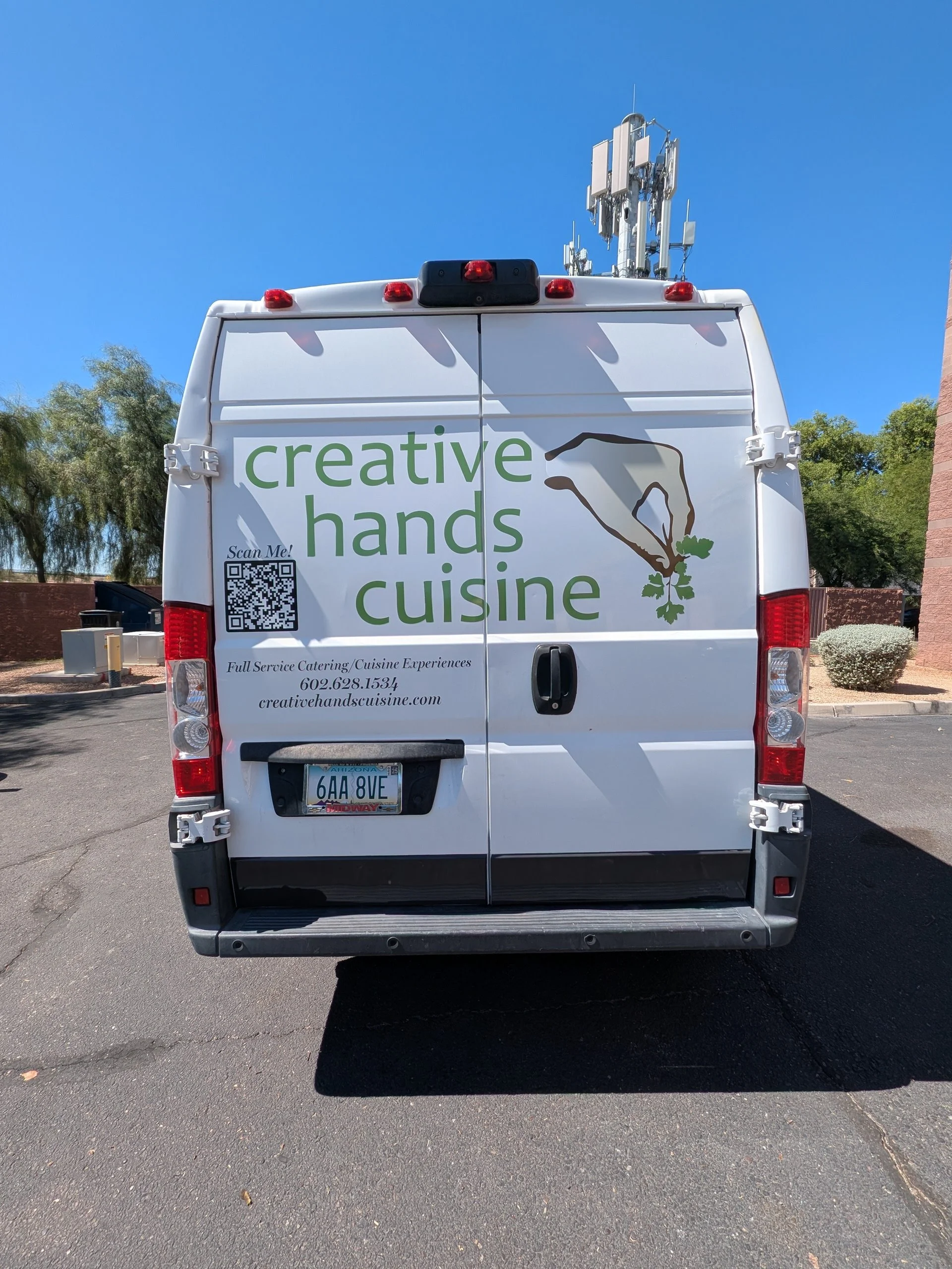 White service van with the logo 'creative hands cuisine' on the back, parked in a lot with trees and a brick wall in the background. The van has a QR code, phone number, and website listed on it.