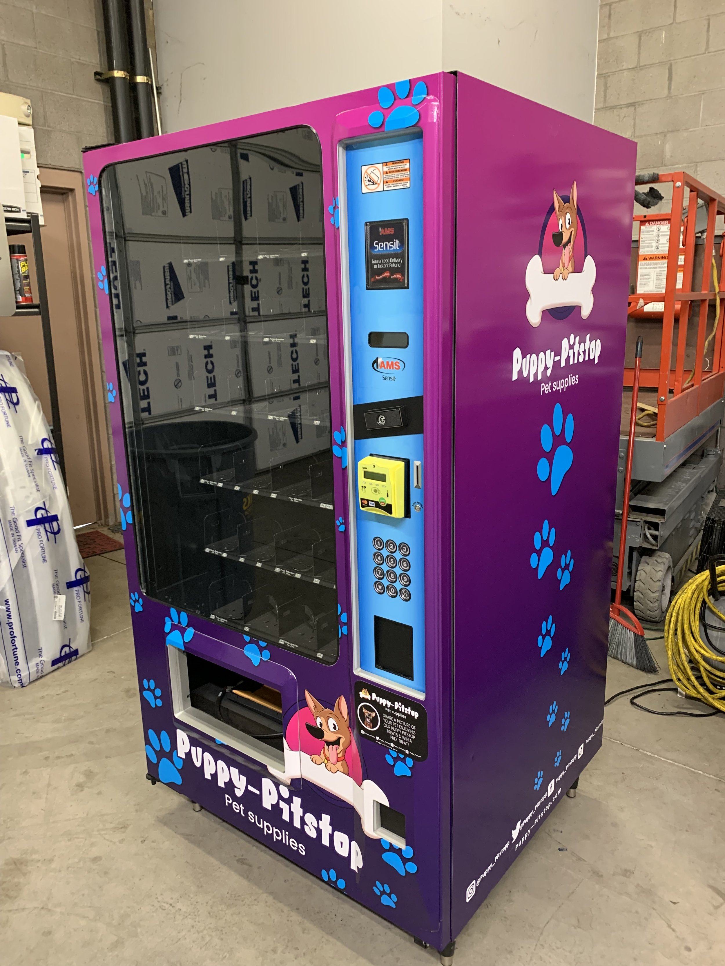 A purple pet supply vending machine with blue paw prints and a cartoon dog holding a bone, labeled "Puppy-Pitstop Pet Supplies." It has a glass door, a control panel, and a yellow payment slot.