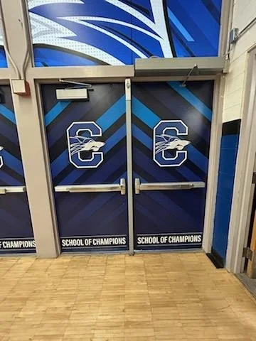 Double doors decorated with the University of Connecticut Huskies logo and the words "School of Champions" at the bottom, located in a sports facility or gym.