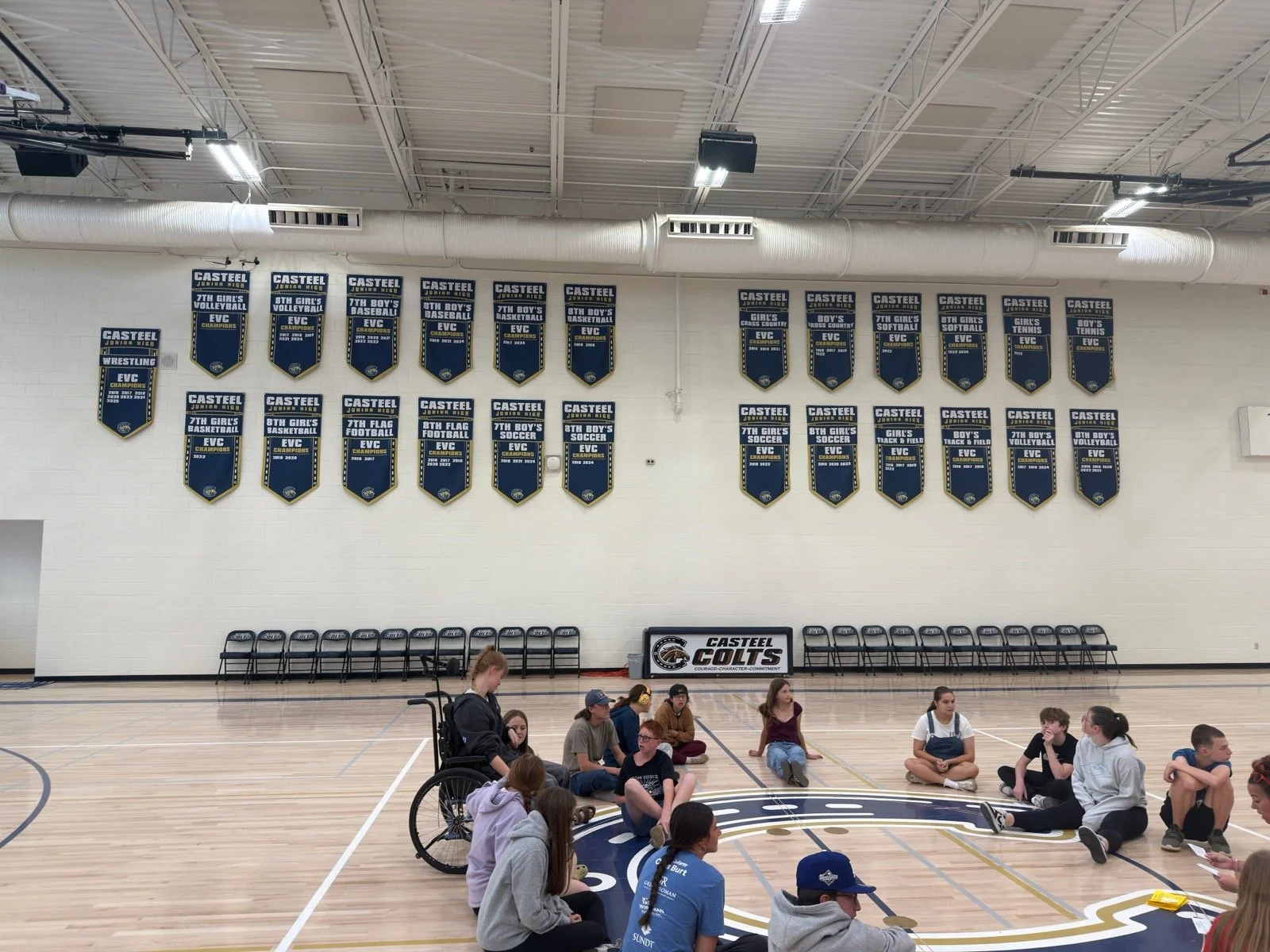 A gymnasium with banners on the wall showcasing various sports achievements of Casteel Junior High School. There are students sitting on the floor in the center of the gym, some in groups, with a wheelchair visible among them. Several chairs are line