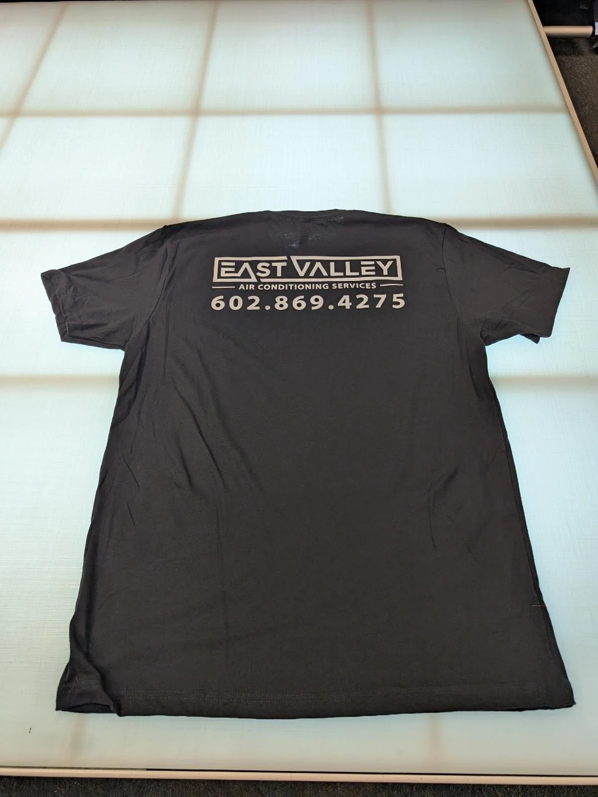 Black T-shirt with white text for East Valley Air Conditioning Services and a phone number, laid flat on a white surface.