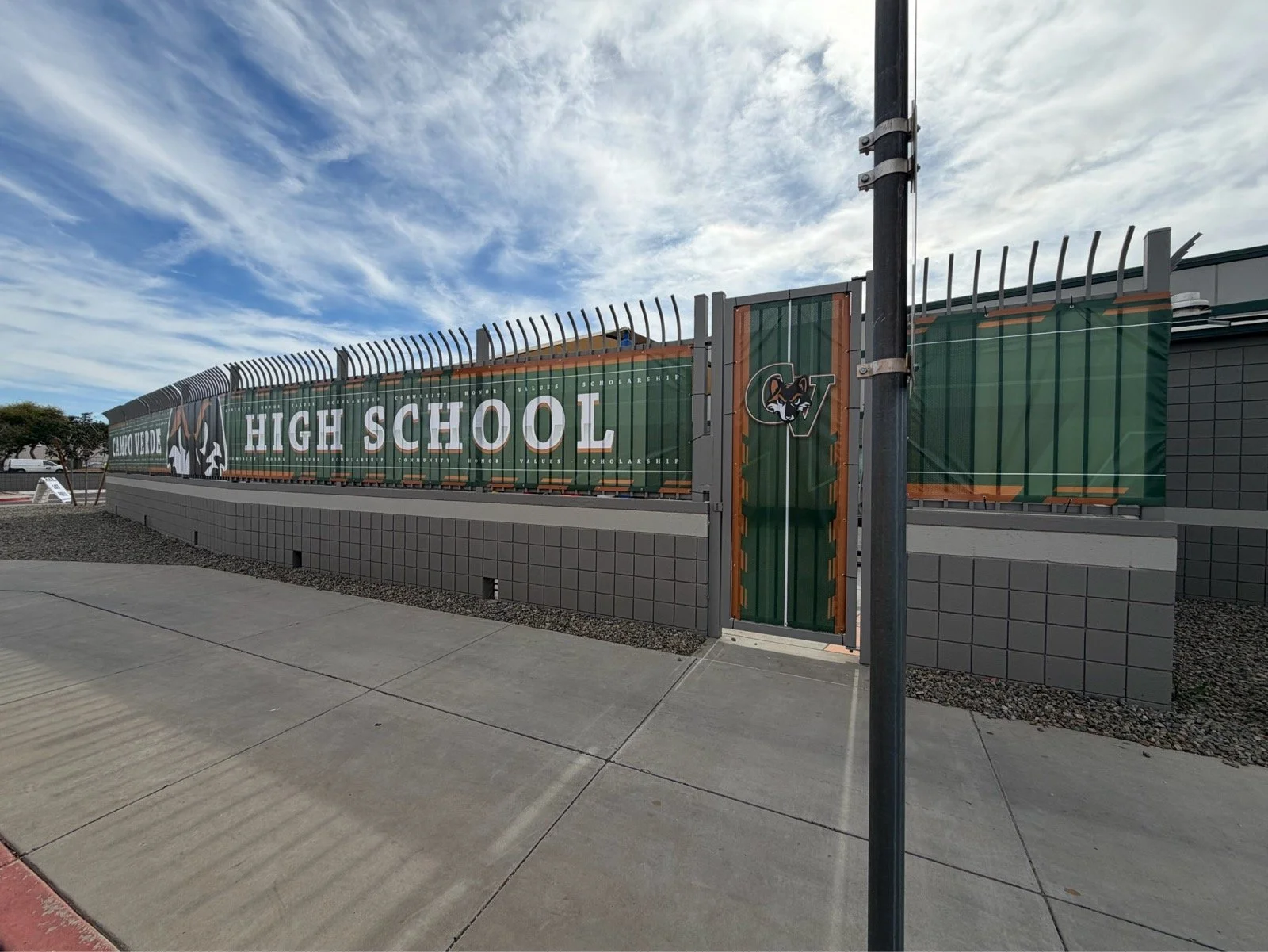 School entrance with a sign that reads 'High School' and a logo of a animal with sharp features, possibly a fox or wolf, behind a tall fence with a gate.