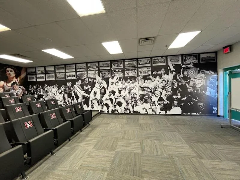 A large black-and-white mural depicting a historic sports team or event on an interior wall in a room with black seats and a green door.