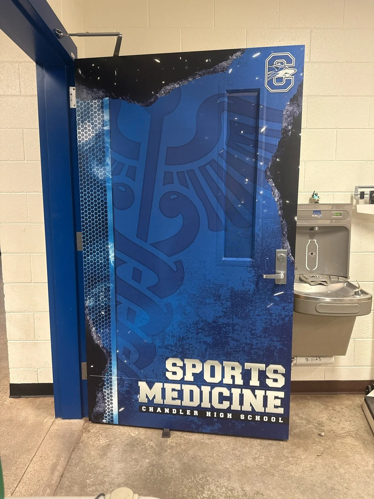 A vending machine decorated with sports-themed graphics for Chandler High School Sports Medicine, featuring a large logo at the top and the words "Sports Medicine" at the bottom. There is a water fountain on the wall next to it.