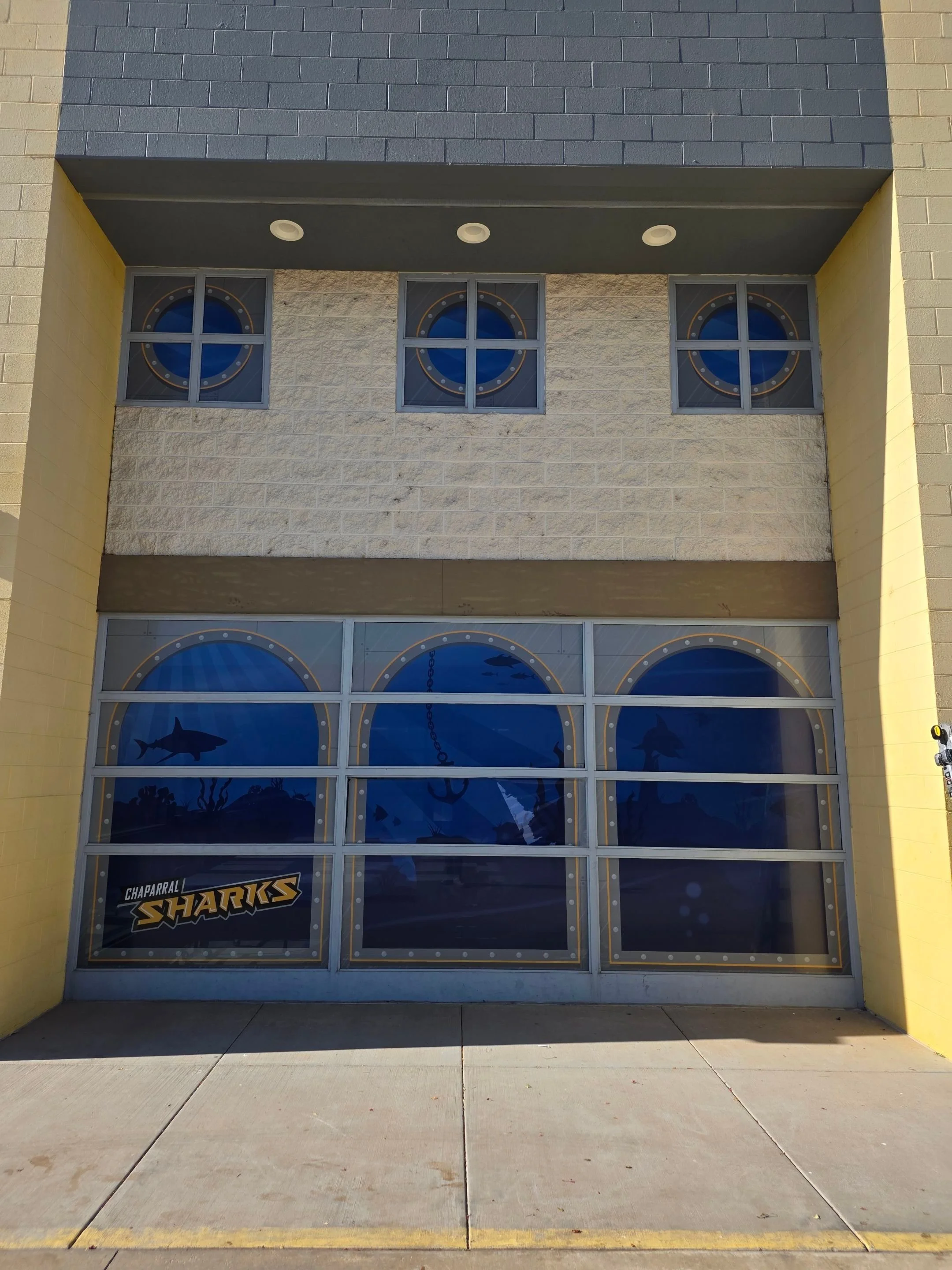 Exterior of a building with a large garage door featuring underwater scene with sharks and seaweed. A sign on the bottom left reads 'Chaparral Sharks'.