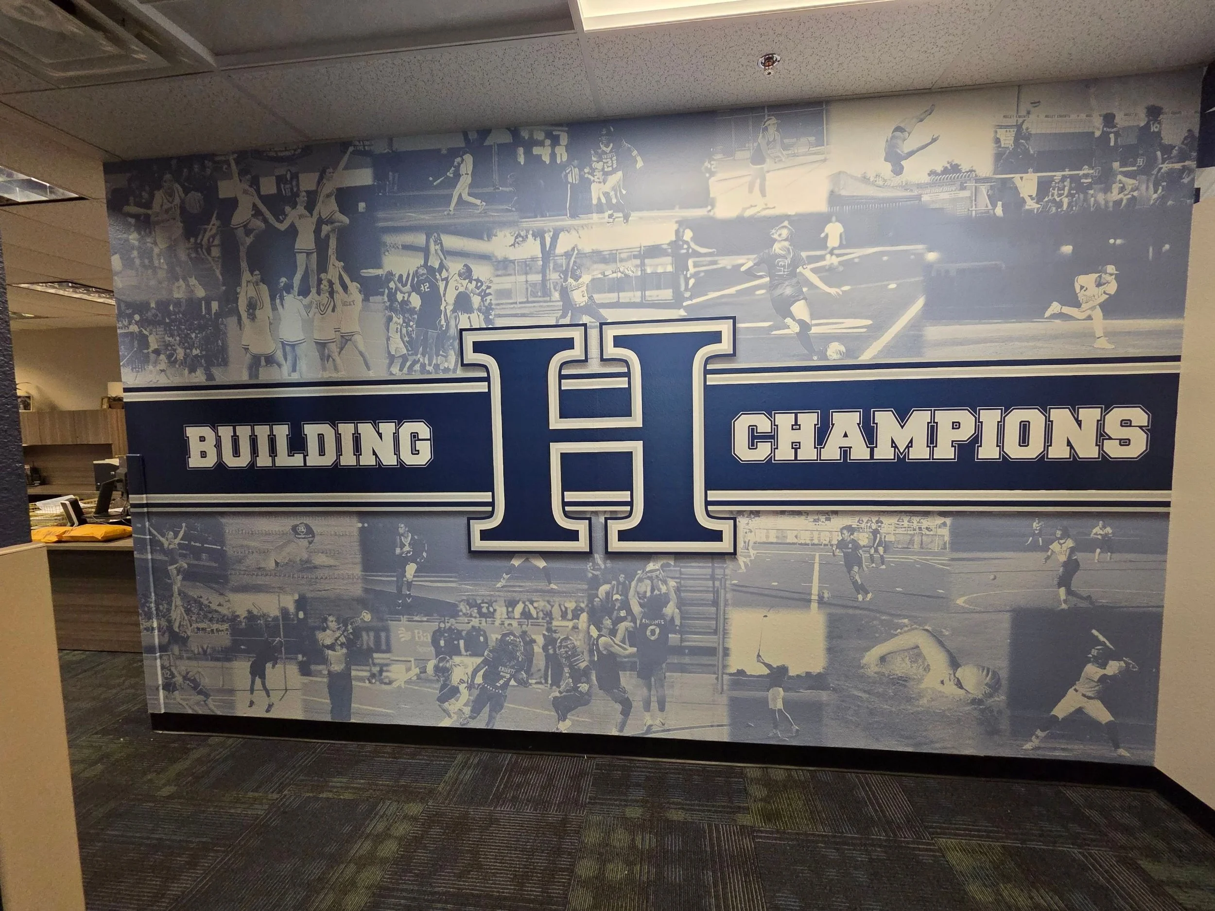 A large wall mural featuring black and white images of basketball players, cheerleaders, and sports scenes with the words 'Building Champions' and a large letter 'H' in the center.