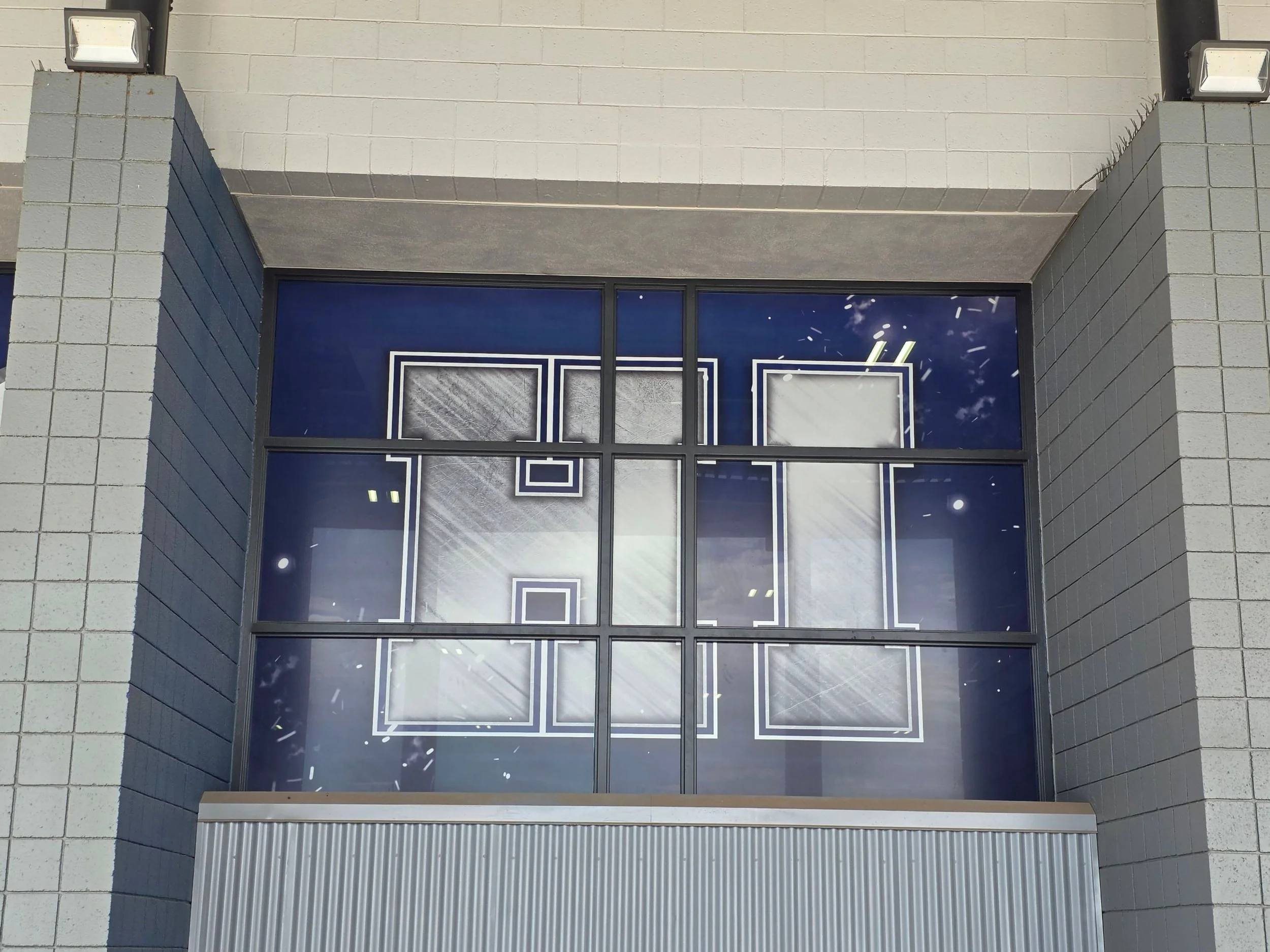 Close-up of a modern building's upper entrance with large glass windows, gray brick walls, and outdoor lighting fixtures.