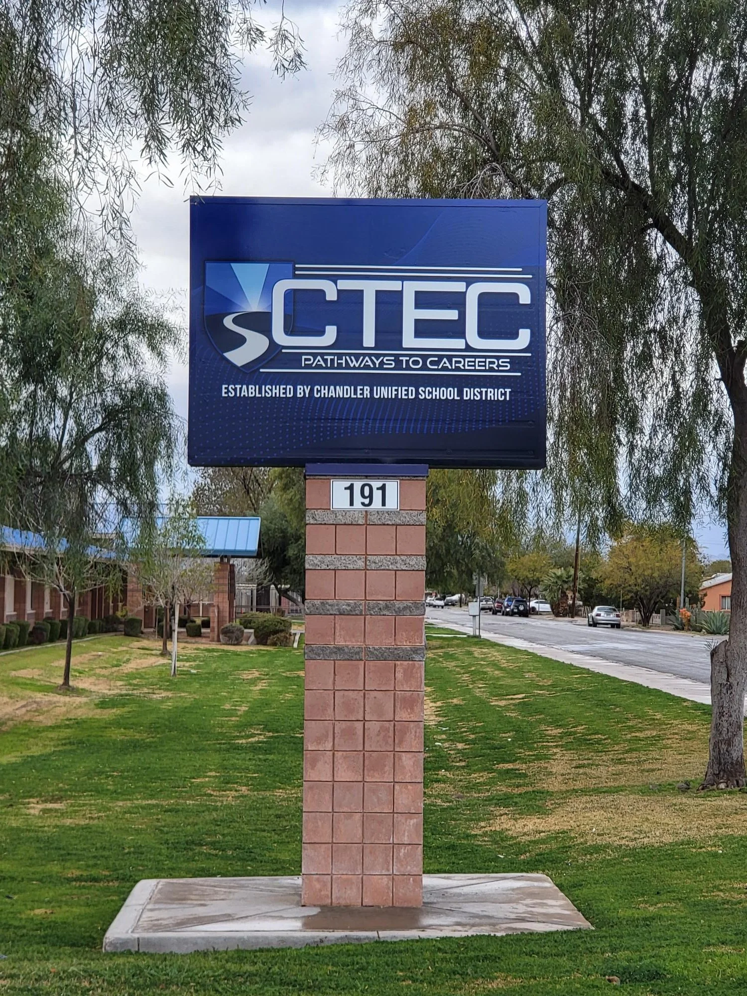 A blue sign with the logo and text for VCTEC, Pathways to Careers, and established by Chandler Unified School District, mounted on a brick and concrete pedestal, with trees, cars, and a street in the background.