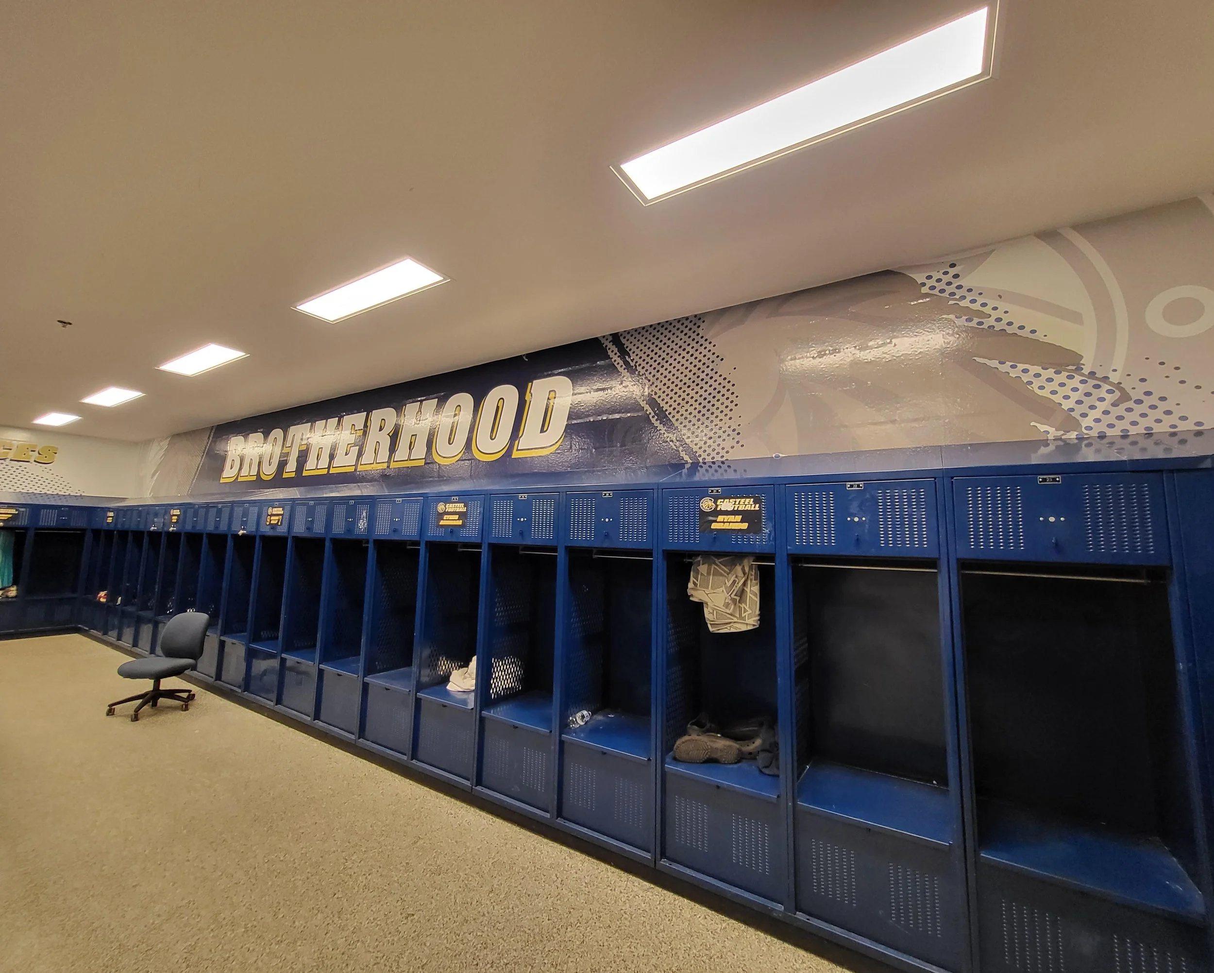 Empty blue lockers under a sign that reads "BROTHERHOOD." There is office chair nearby, shoes on the floor, and fluorescent ceiling lights.