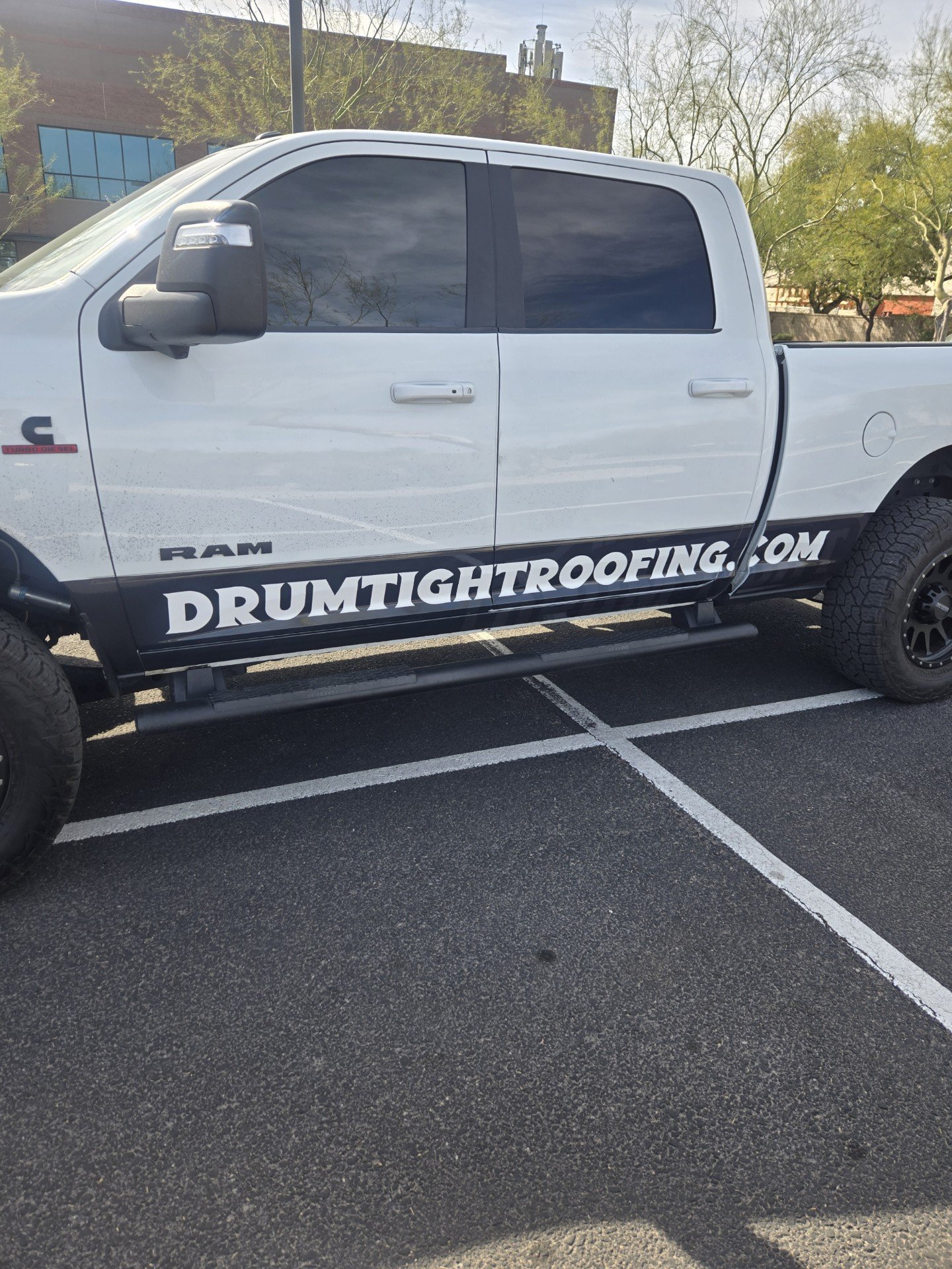White RAM pickup truck parked in an outdoor parking lot with trees and building in the background, advertising for drumtightroofing.com on the side.