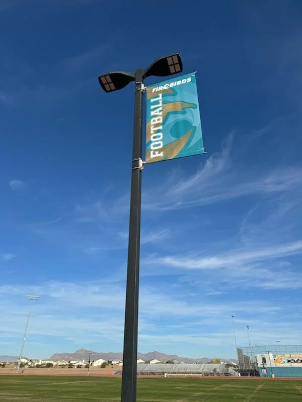 A tall light pole with a banner that says 'FIREBIRDS FOOTBALL' and features a stylized bird logo, set against a blue sky with some clouds, beside a sports field.