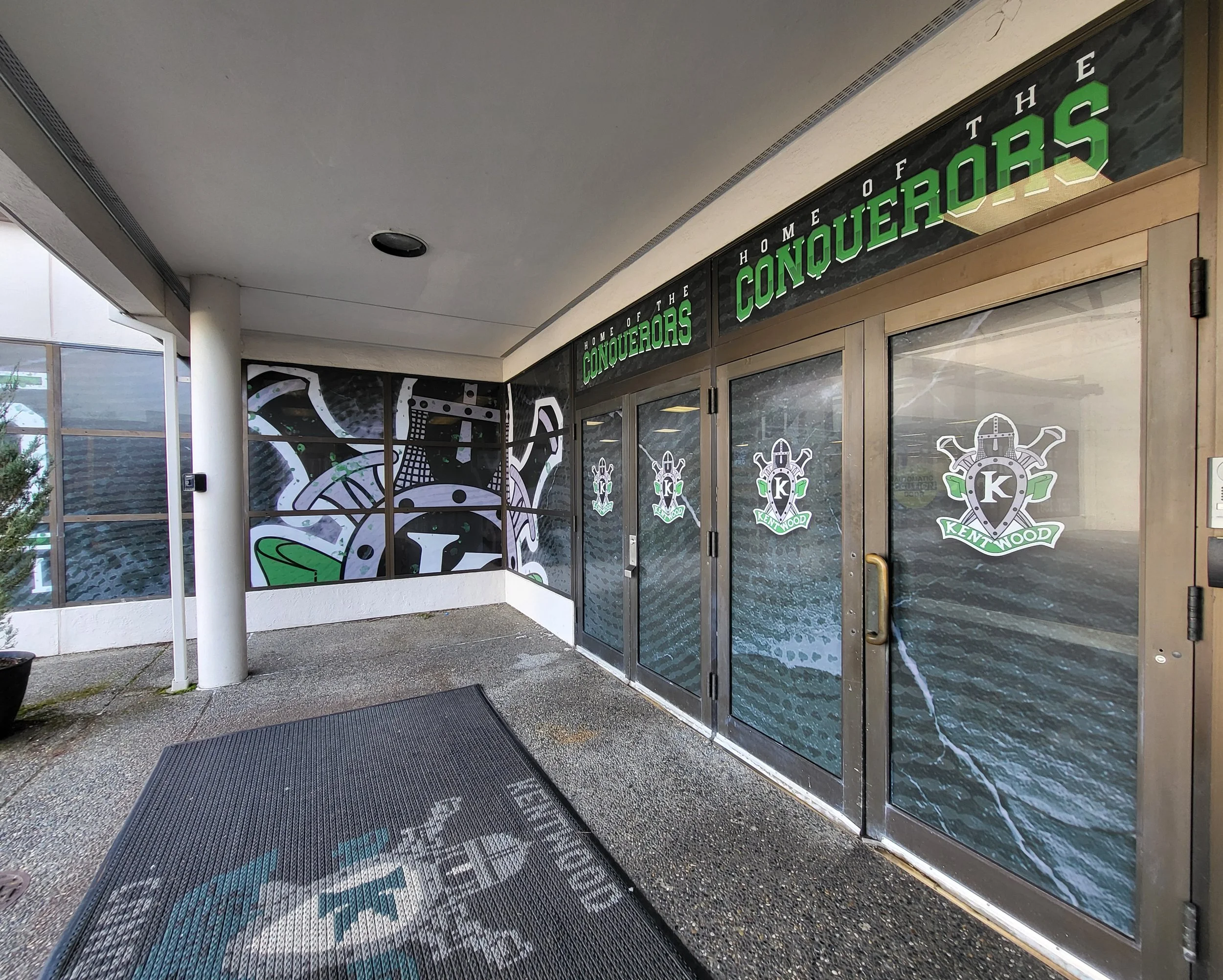Storefront with signs reading 'Home of the Conquerors' in large green letters, decorated with logos of a knight's helmet and tools, and glass doors with decals of various tools.