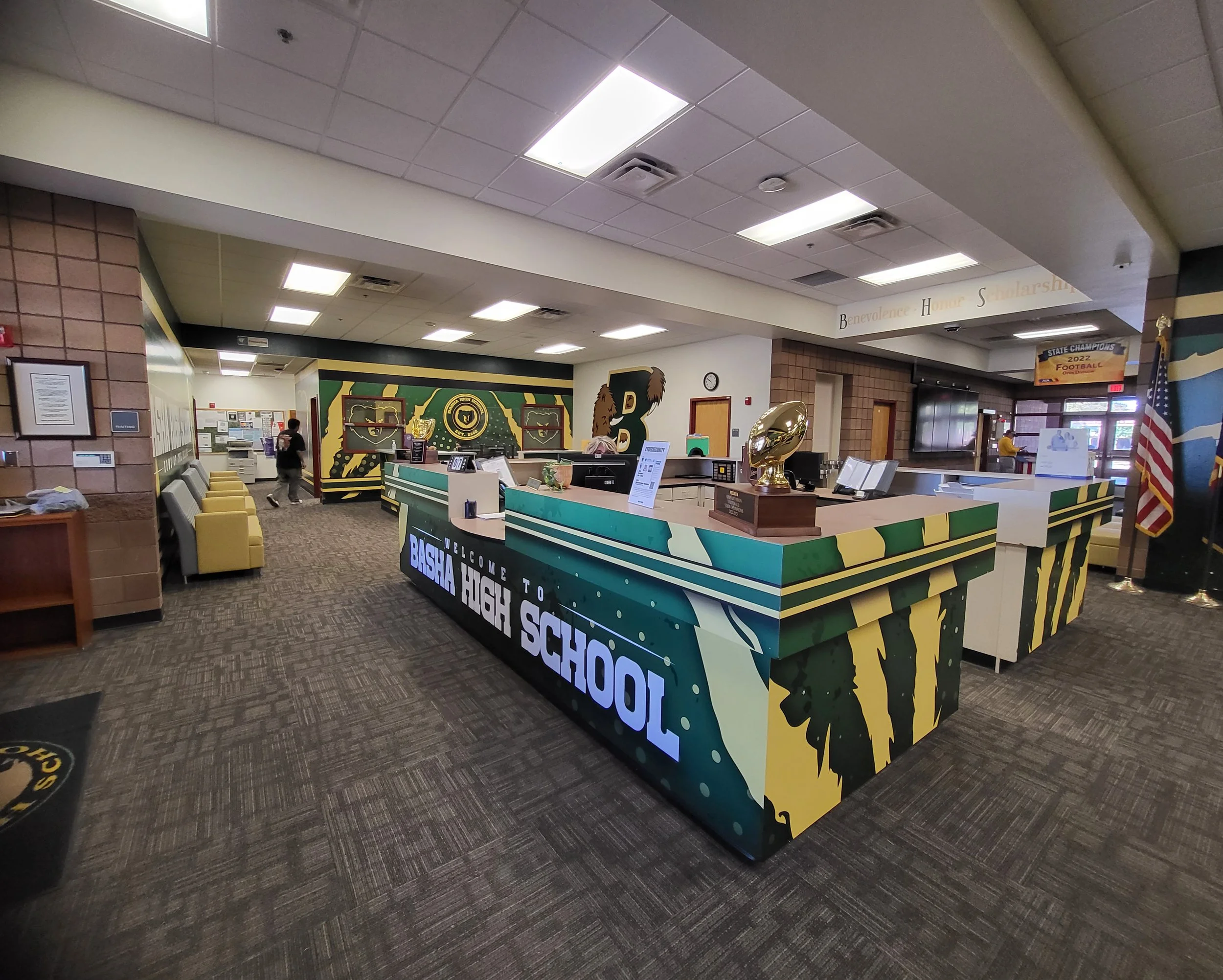 The reception desk at a high school with a sign that says, "Welcome to Bishop High School." The front desk has informational signs, a computer, and a large golden trophy. Behind the desk, there is a colorful mural with the letter "B" and a depiction 